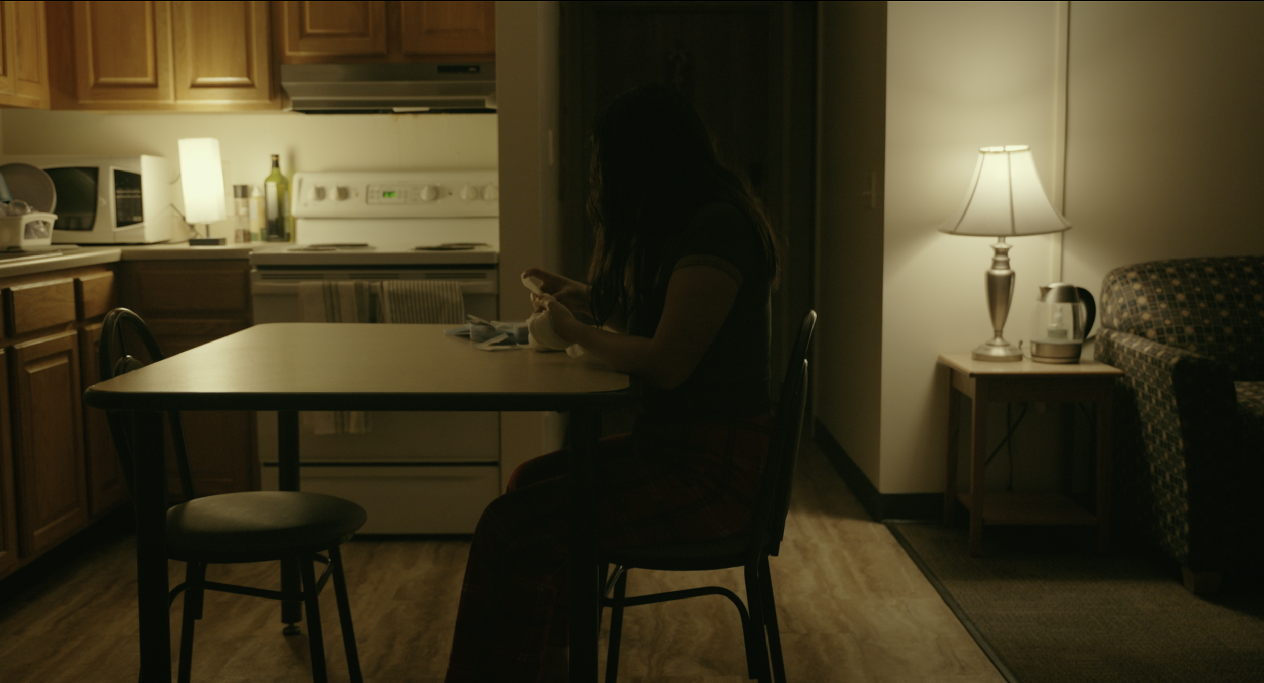 A woman with long dark hair sitting at a kitchen table in a dimly lit room, using a smartphone. The kitchen background has wooden cabinets, a stove, and various appliances. A table lamp on a side table provides warm lighting.