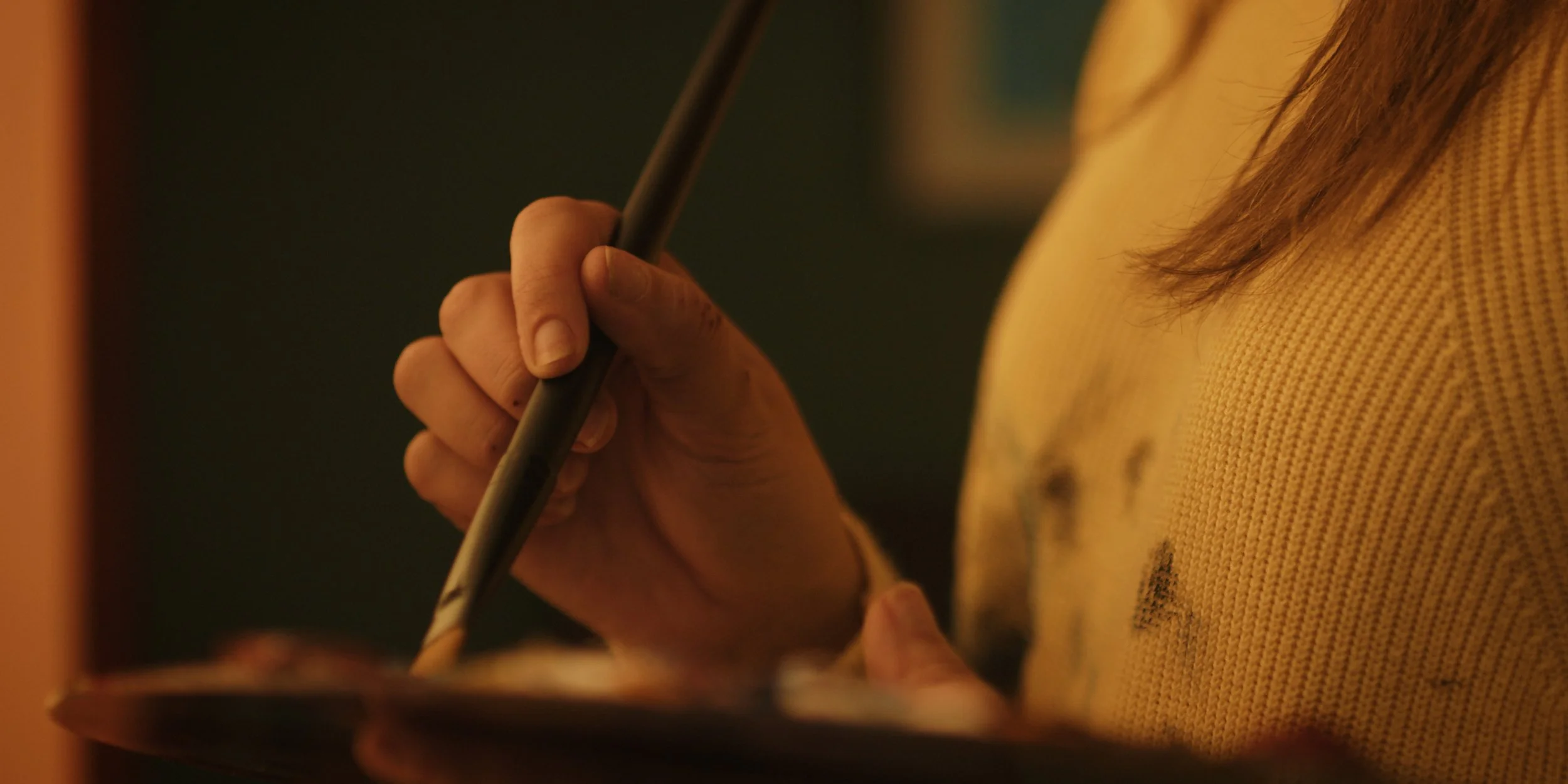 Close-up of a person's hand holding a paintbrush, painting on a surface, with part of their yellow sweater visible.