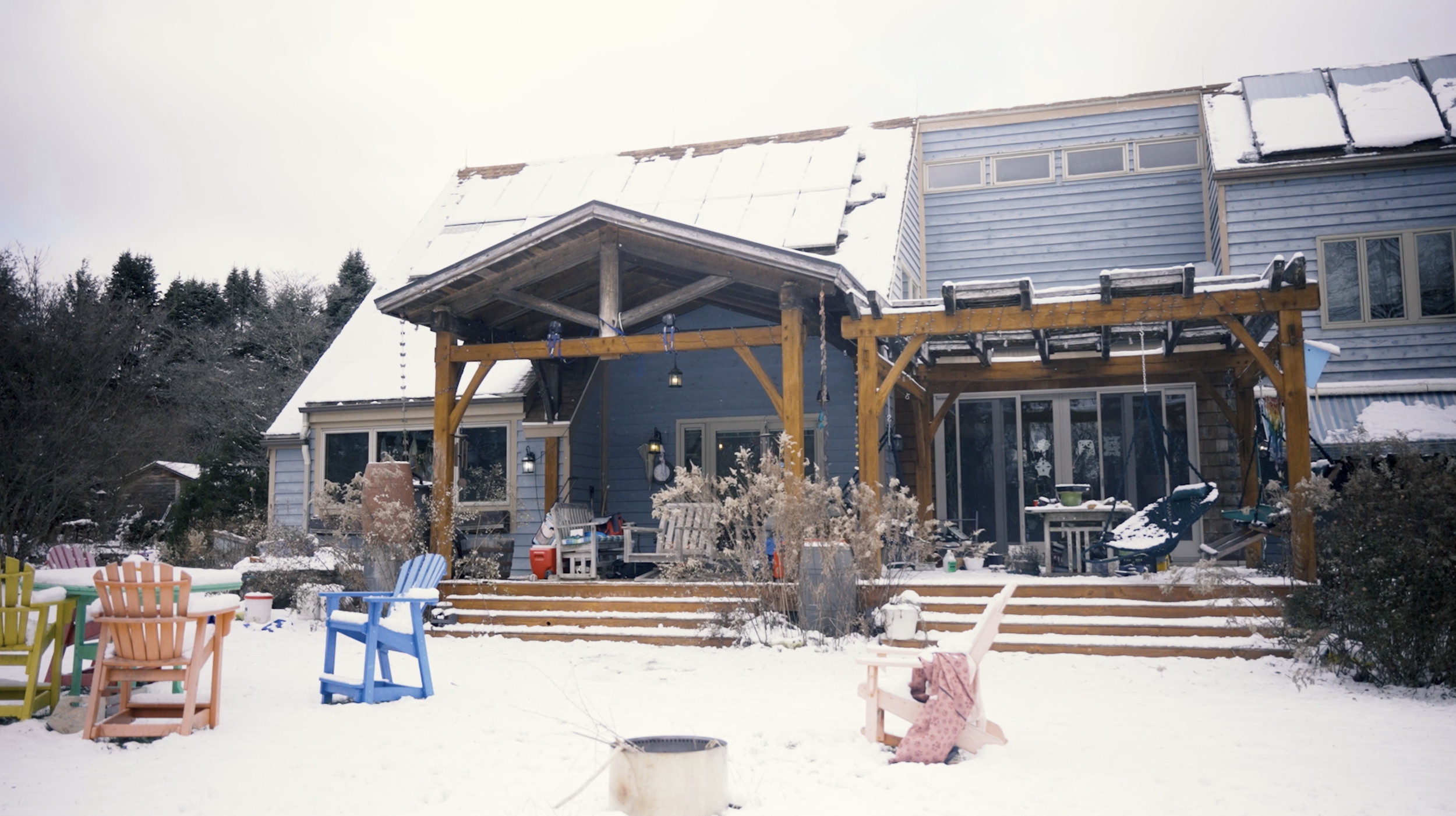 Backyard scene with snow-covered ground, colorful chairs, a large house with a second-story porch, and trees in the background.