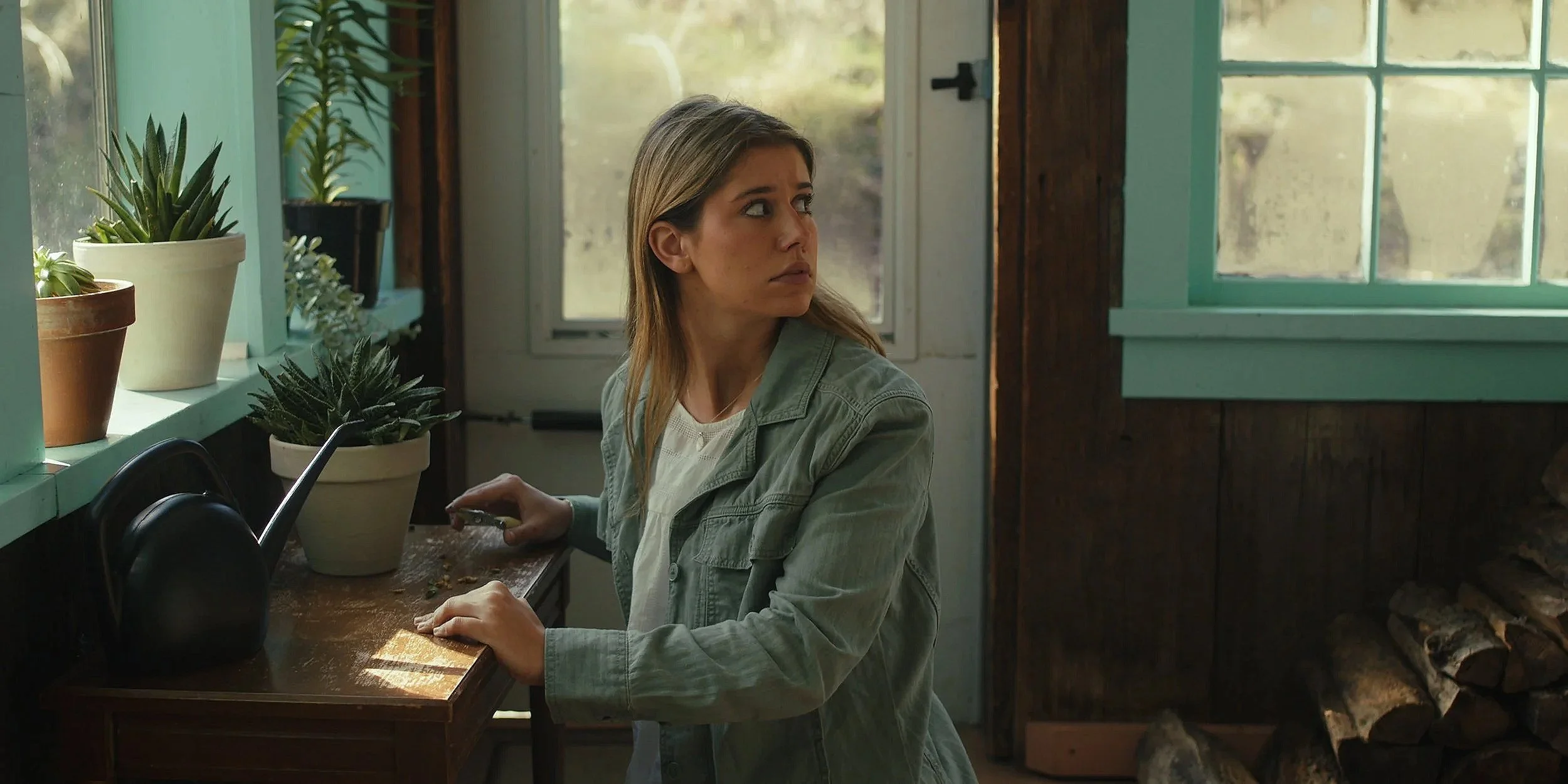 A woman with long hair sitting at a wooden table inside a rustic room with large window, potted plants on the windowsill, and stacks of firewood behind her. She is looking to her left with a neutral expression, holding a device in her right hand.