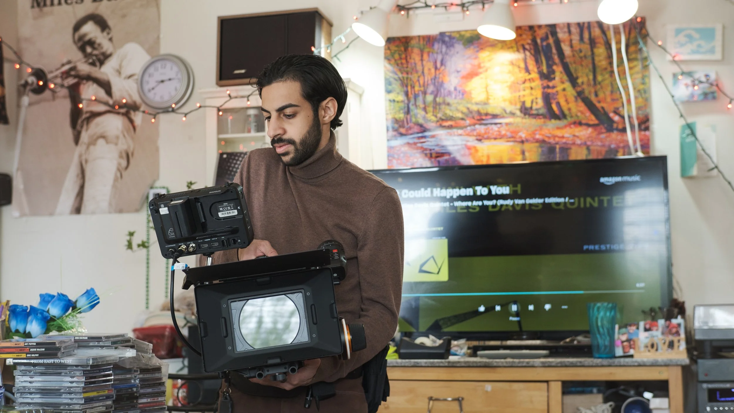 A man with dark hair and a beard, wearing a brown turtleneck, standing in a room decorated with artwork, string lights, and a vintage poster of Miles Davis. He is holding a camera or video equipment, focusing on it. Behind him, there is a TV screen displaying text and images, and on the table are stacks of DVDs, books, and decorative items.