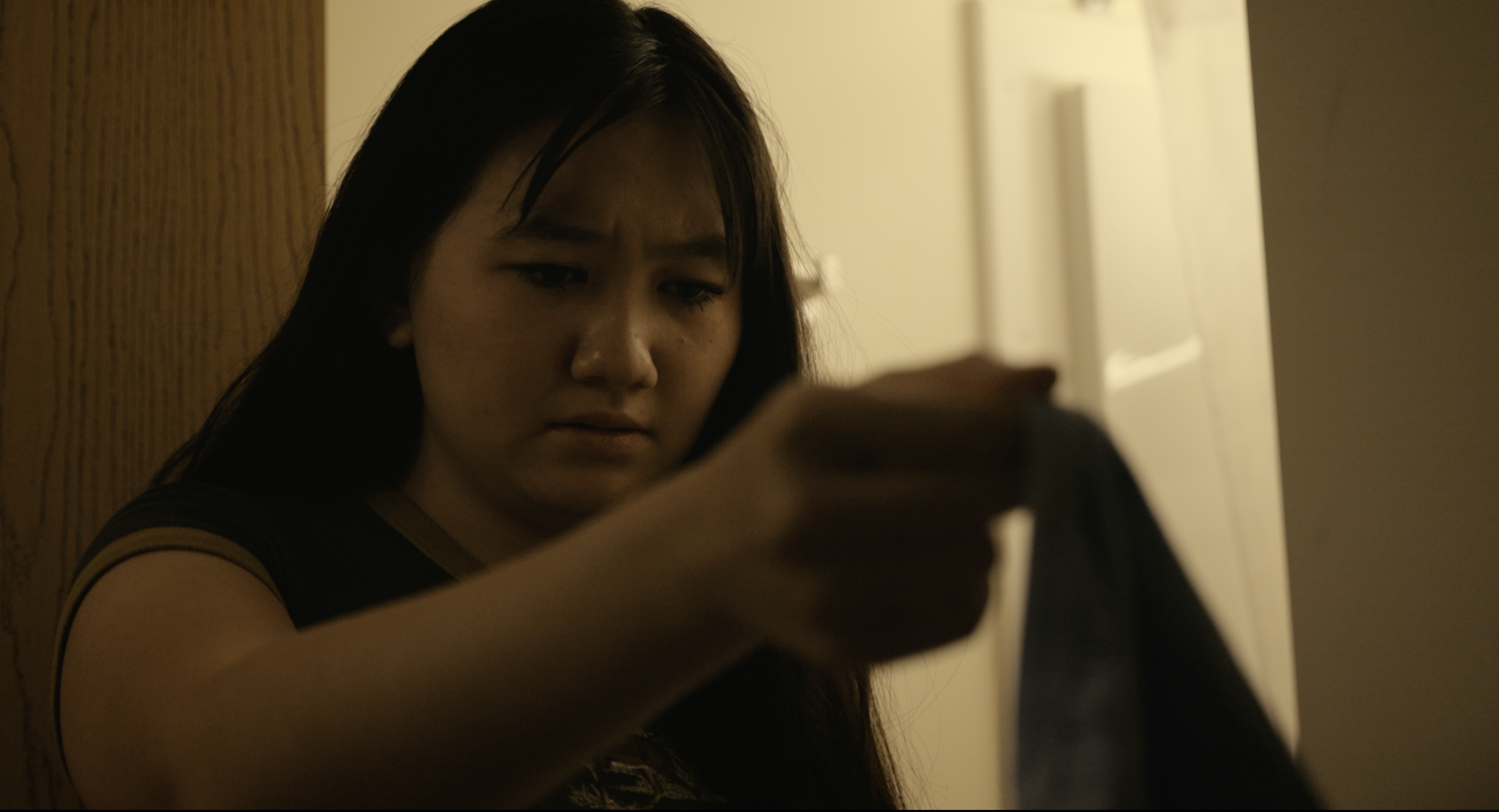 A young woman with long dark hair, wearing a dark shirt with yellow trim, is looking down and appears to be focused on something she is holding or working on, against a background of a wooden wall and a doorway.
