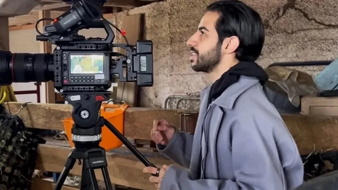 A man with dark hair and a beard filming with a professional video camera in a workshop or garage setting.