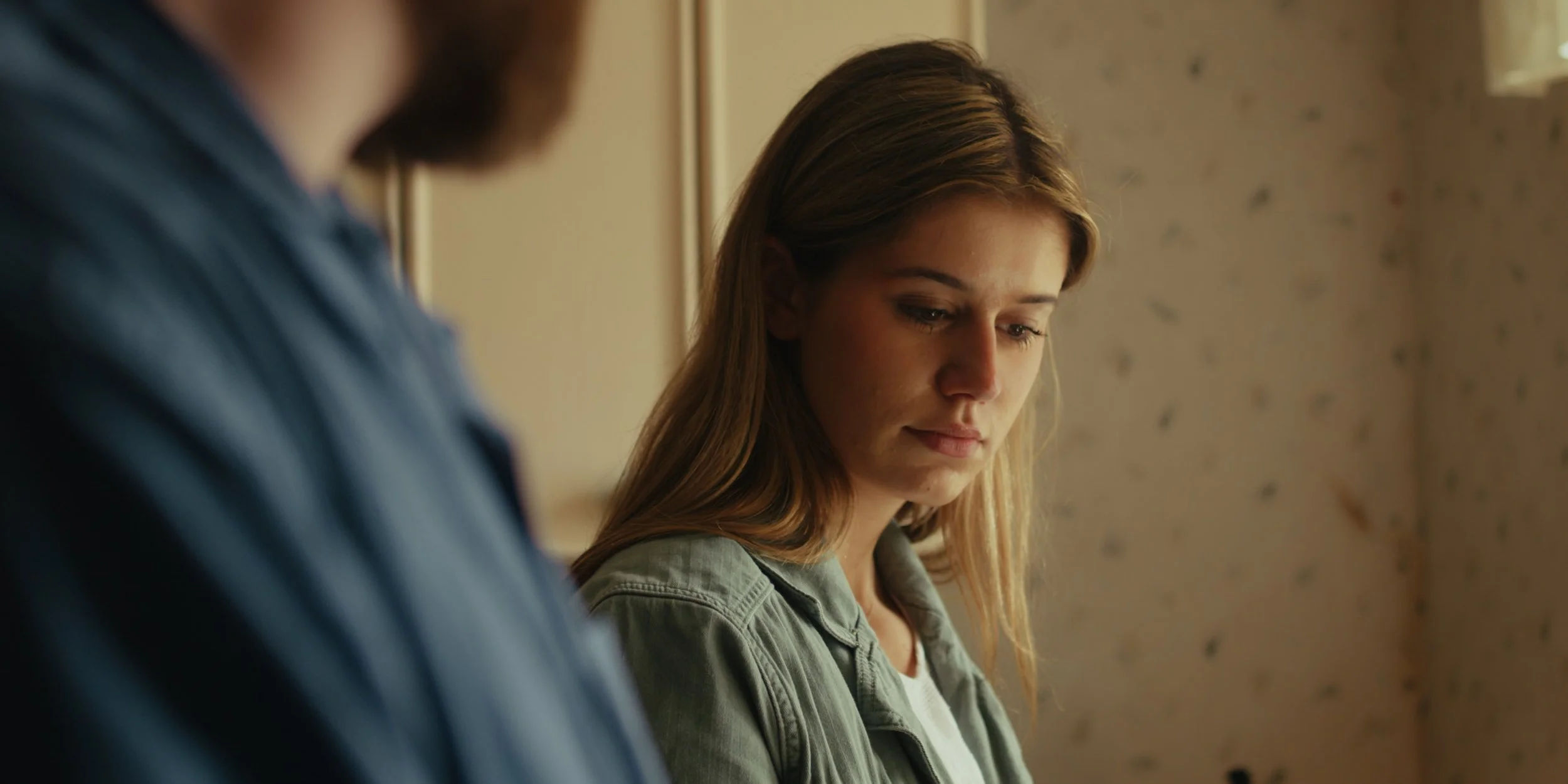 A young woman with light brown hair looking down, with part of a person in a blue shirt visible in the foreground.