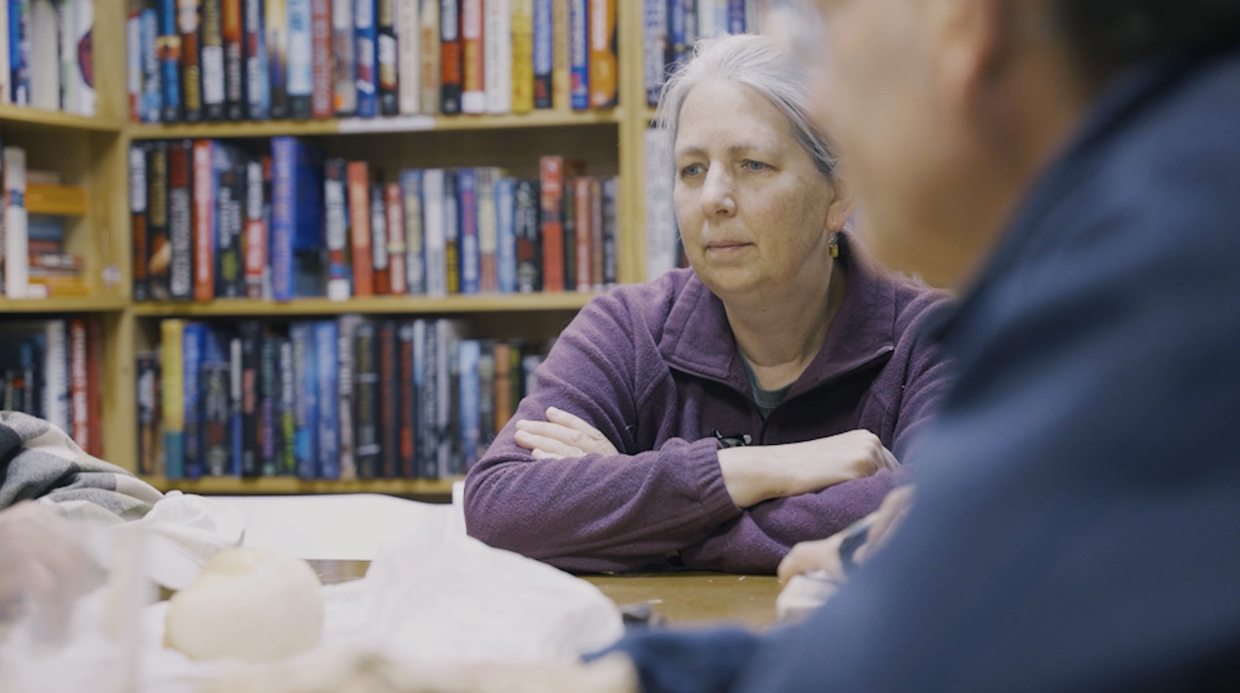 Two women sitting at a table in a library, engaging in conversation, with bookshelves in the background.