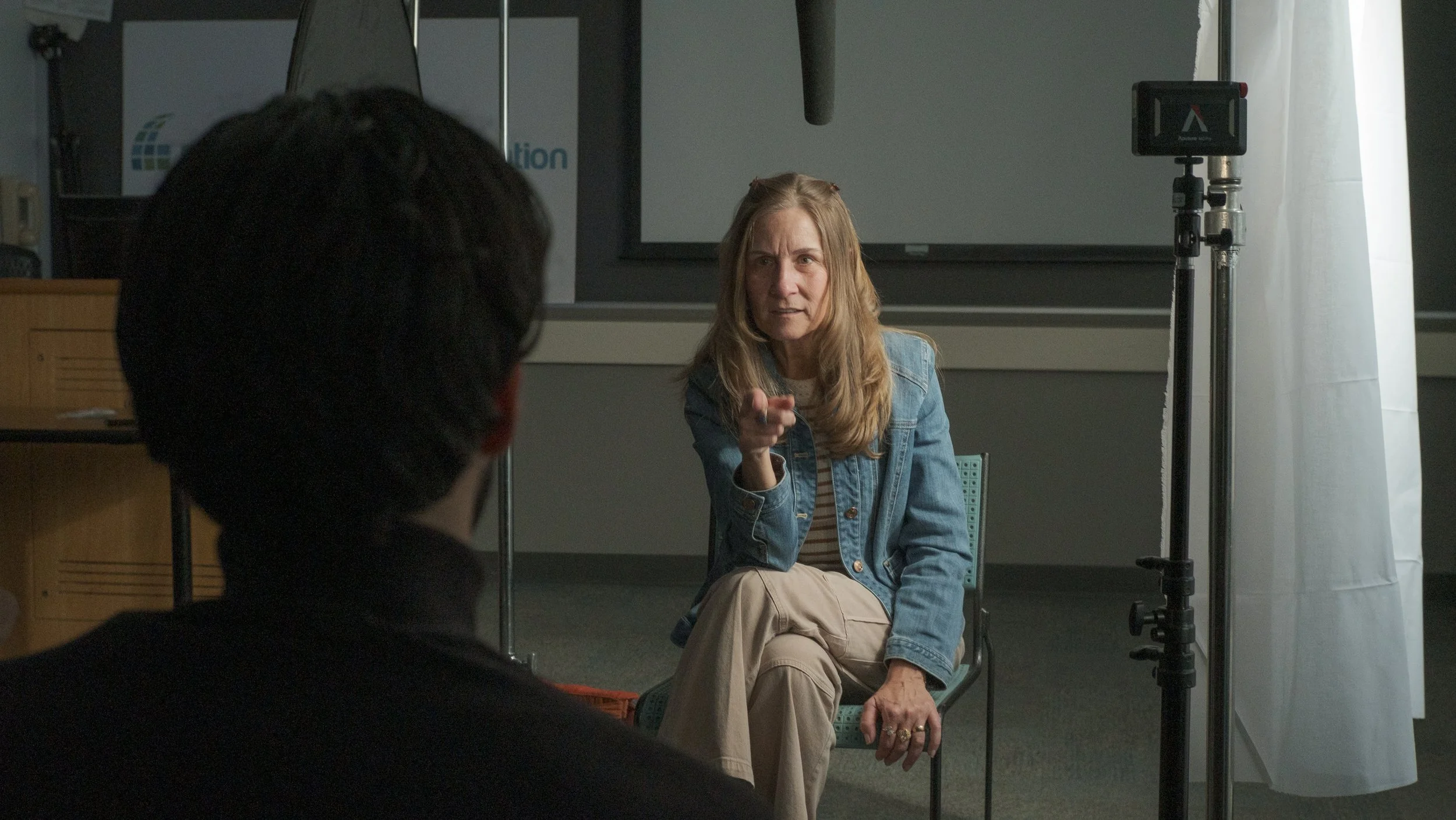 A woman with long blonde hair wearing a denim jacket and tan pants, sitting and pointing while speaking during an interview or conversation in a studio.