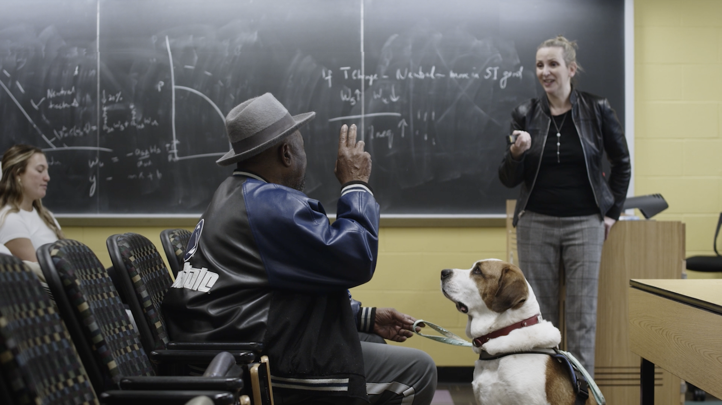 A classroom scene with a woman teaching and three students, including a man with a hat and a dog, seated at desks in front of a chalkboard filled with mathematical notes.