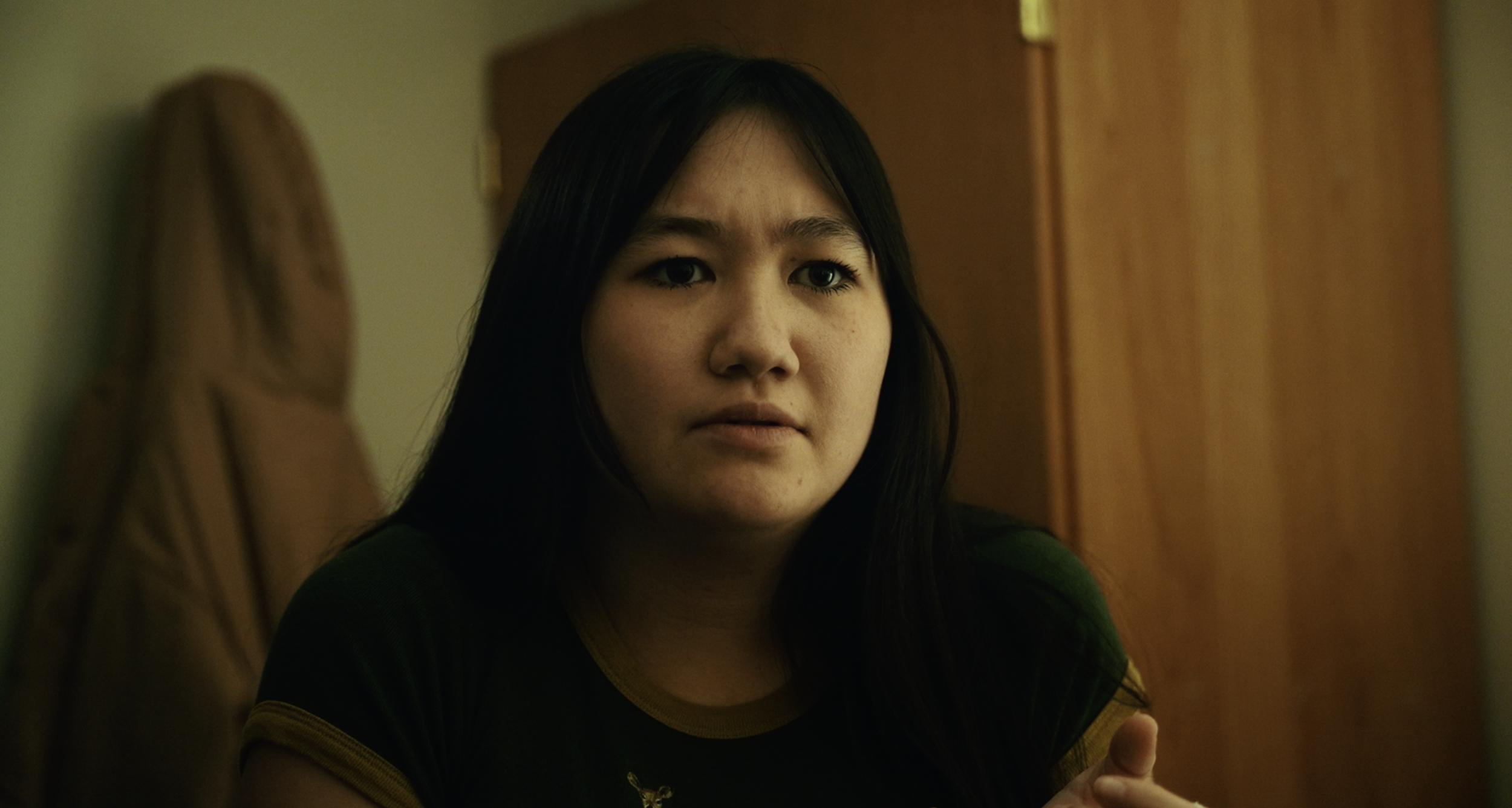 A woman with long black hair looks contemplative, sitting indoors near a wooden door, wearing a black shirt with yellow trim.
