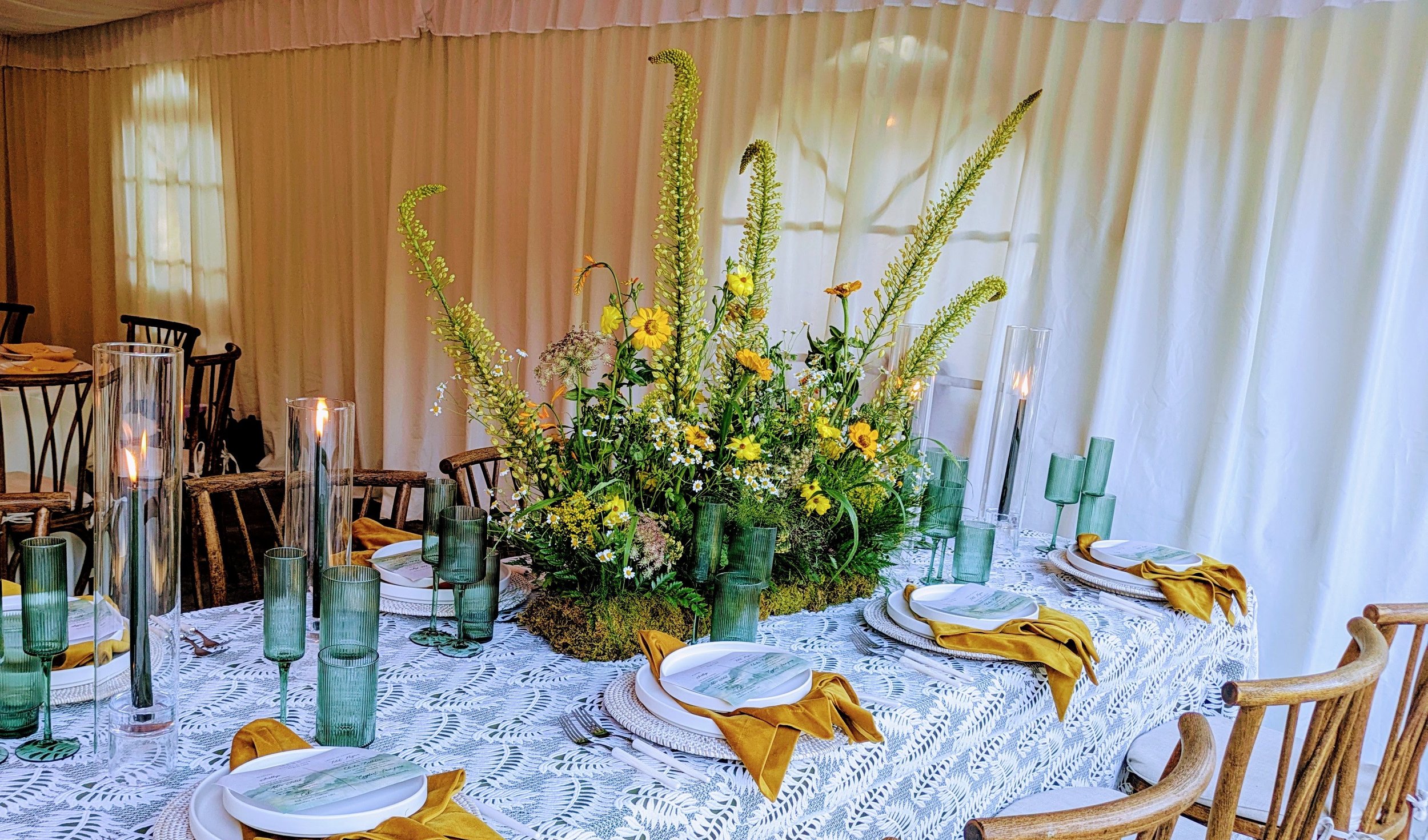 A decorated dining table with a large flower arrangement centerpiece, surrounded by green glassware, white plates with gold napkins, and wooden chairs, set against a backdrop of closing white curtains.