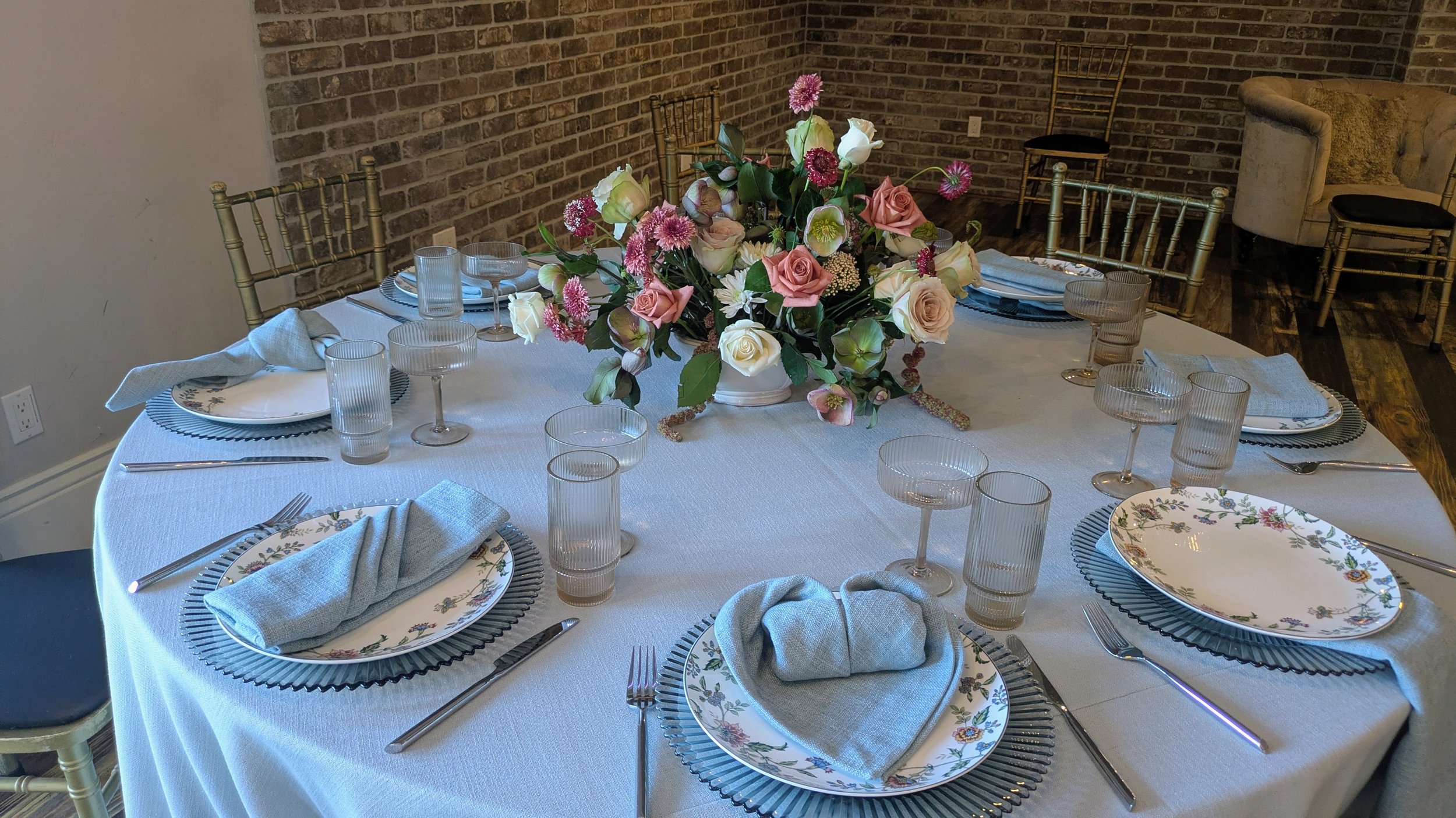A dining table set for a meal with a white tablecloth, floral-patterned plates, silverware, cloth napkins, and glassware. The centerpiece is a large bouquet of pink, white, and green flowers. Surrounding the table are gold-colored chairs and a brick wall in the background.