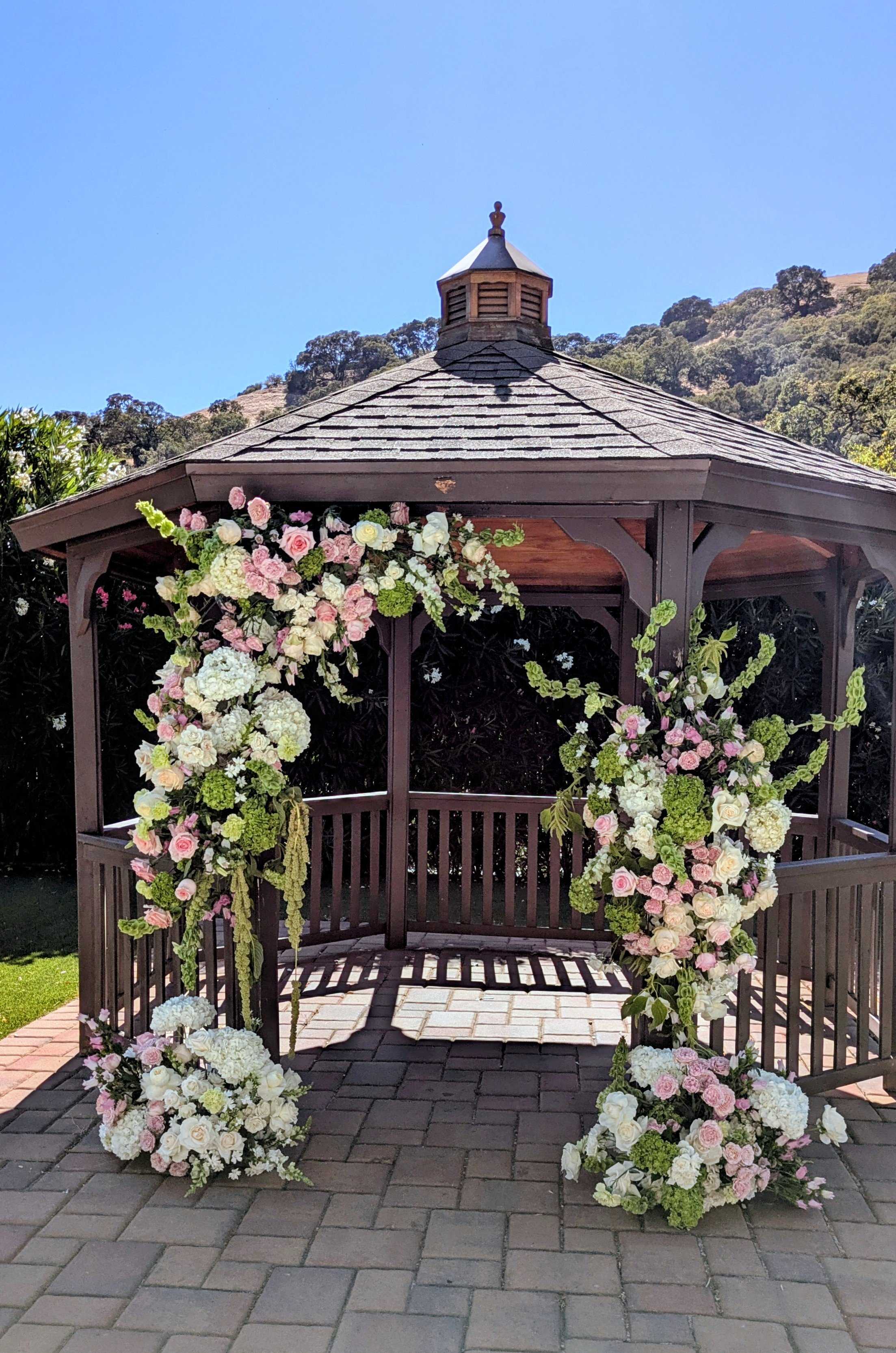 A wooden gazebo decorated with pink and white flowers, situated outdoors on a brick-paved area with a backdrop of green trees and a hillside under a clear blue sky.