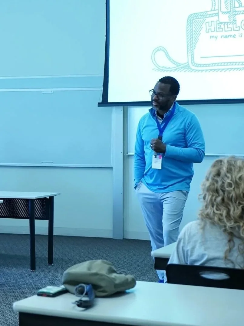 A man stands in front of a classroom giving a presentation, with a slide projected on a screen behind him. The slide has a doodle of a bag and the word 'HELLO' on it. He wears glasses, a blue shirt, and a lanyard. Students sit at desks, one with a backpack and phone on it.