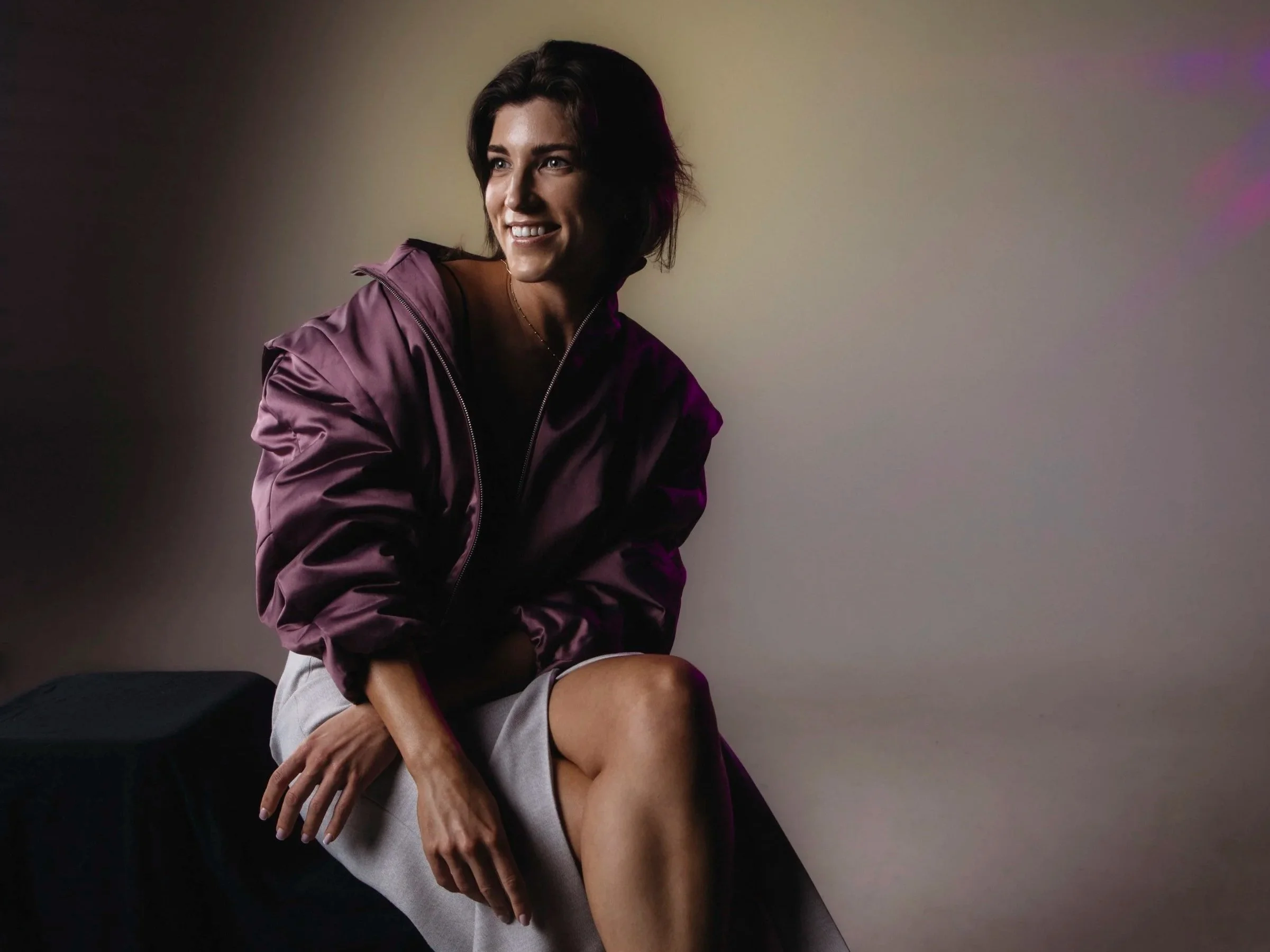 A woman with short dark hair smiling, wearing a purple jacket and light-colored shorts, sitting on a man's lap against a neutral background.