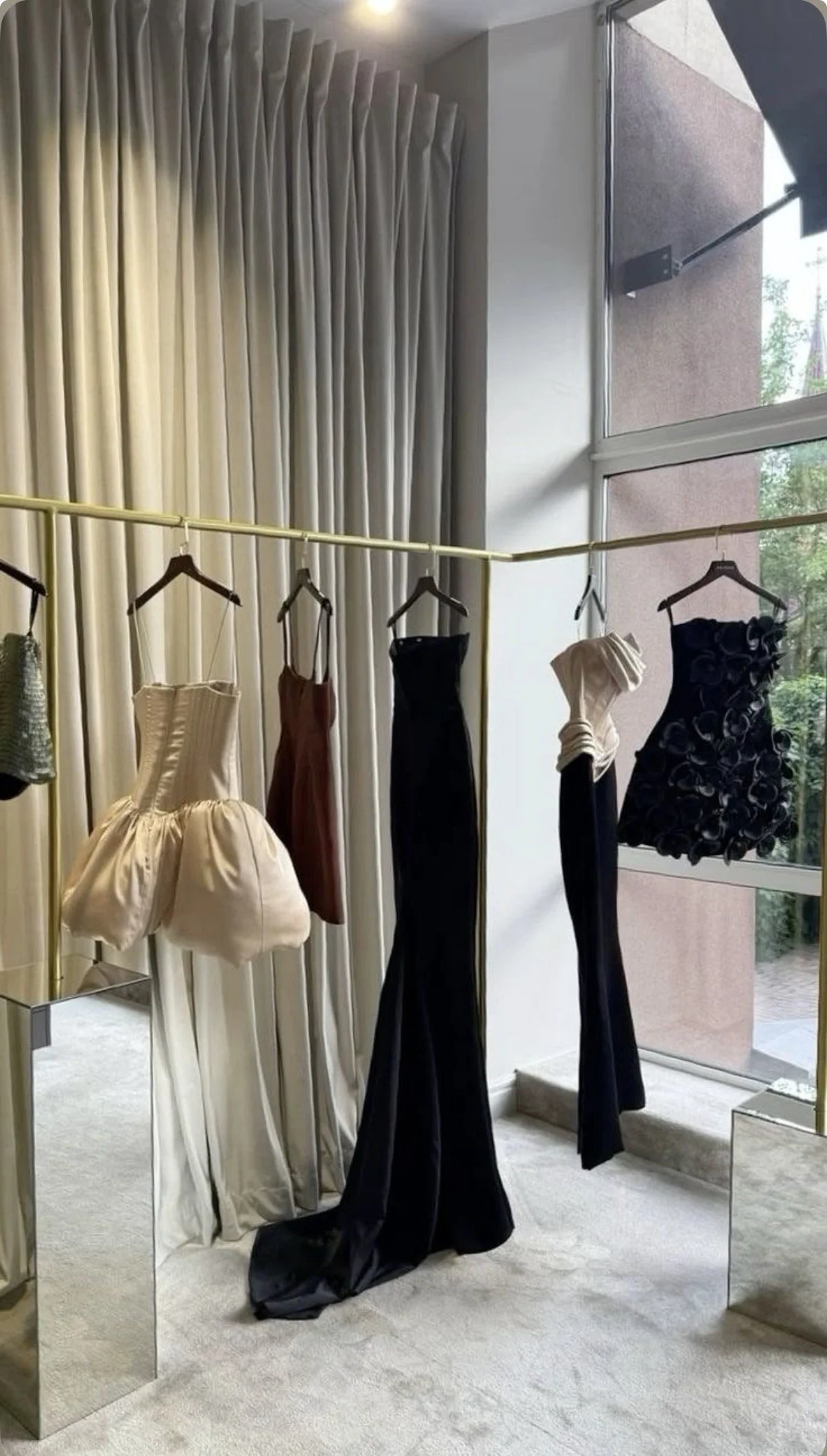 Elegant dresses hanging on a clothing rack in a room with floor-to-ceiling beige curtains and large windows. The dresses include a cream-colored ball gown, a brown dress, a black dress, and a black dress with floral details. A mirror reflects the dresses and the room.