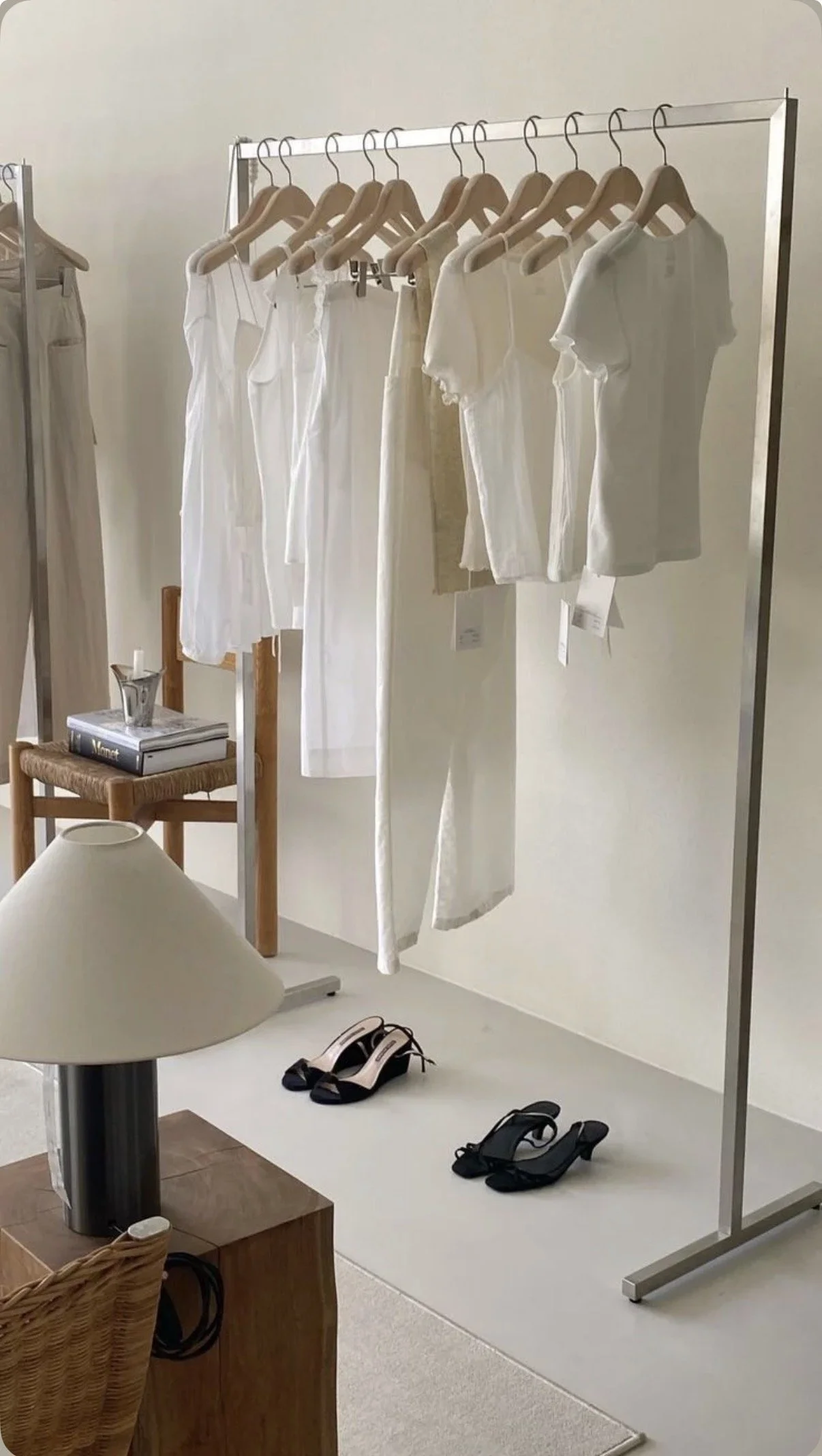 White clothing hanging on a rack in a showroom or boutique, with shoes on the floor below and a table with books and a small plant nearby.