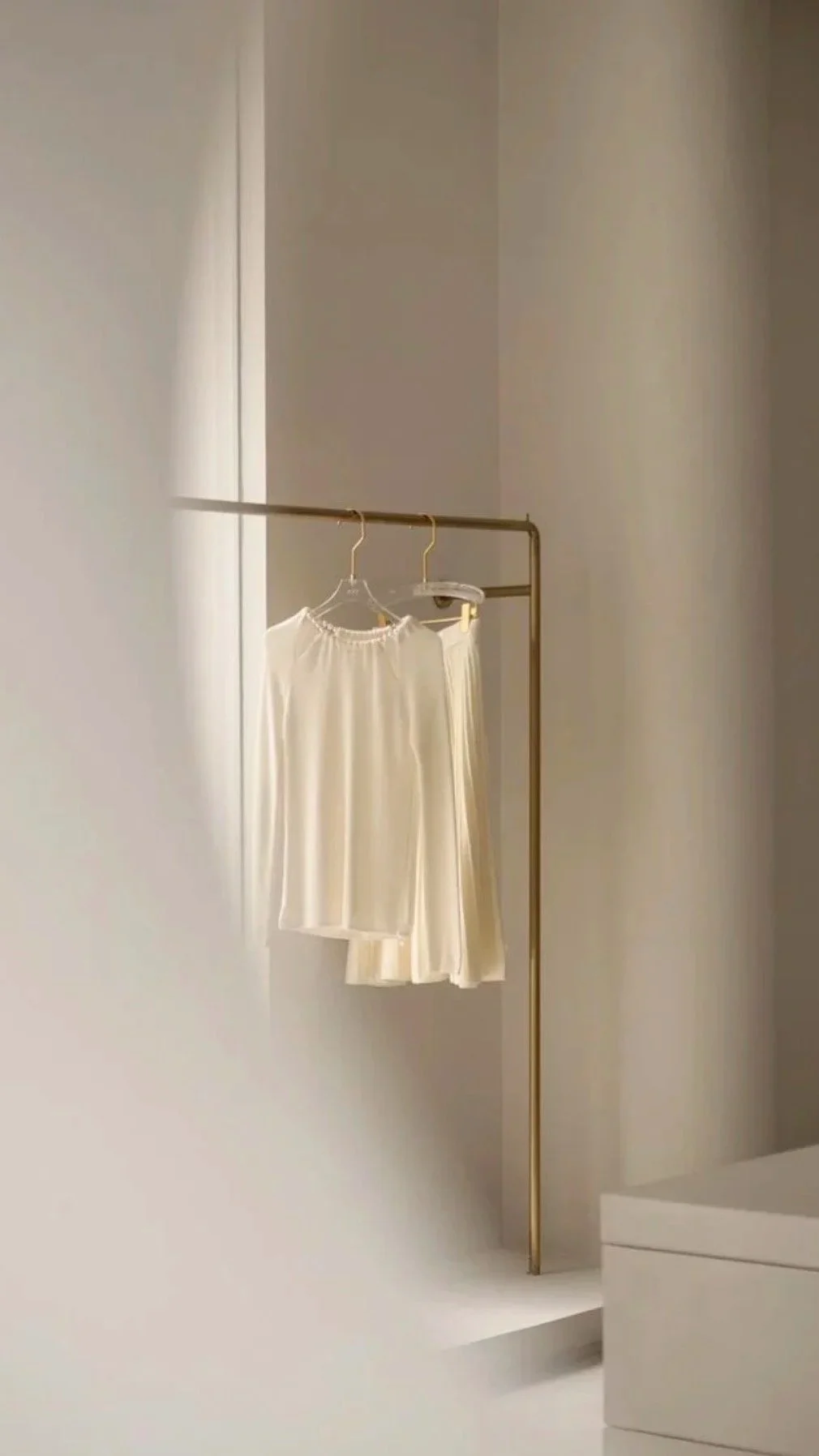 A minimalist clothing display with a gold clothing rack, holding a cream-colored dress and skirt, set against a neutral wall.