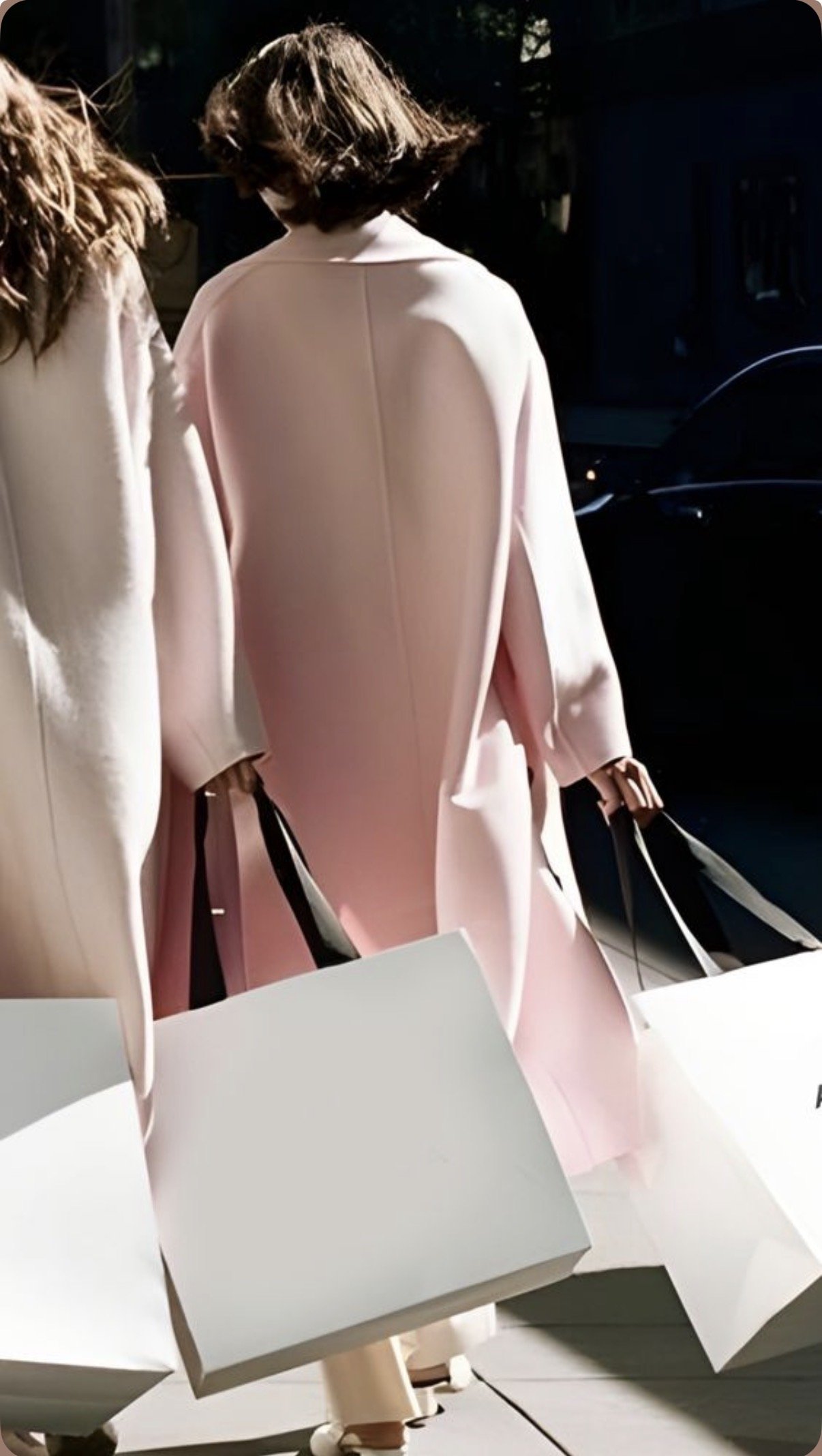 Two women wearing light-colored coats carrying shopping bags walk outdoors, with sunlight casting shadows on their backs.
