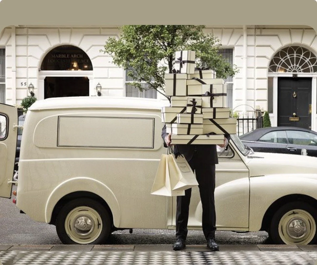 Person standing next to a vintage cream-colored van, holding several gift boxes wrapped in beige paper with black ribbons, and shopping bags, on a city street lined with white buildings.