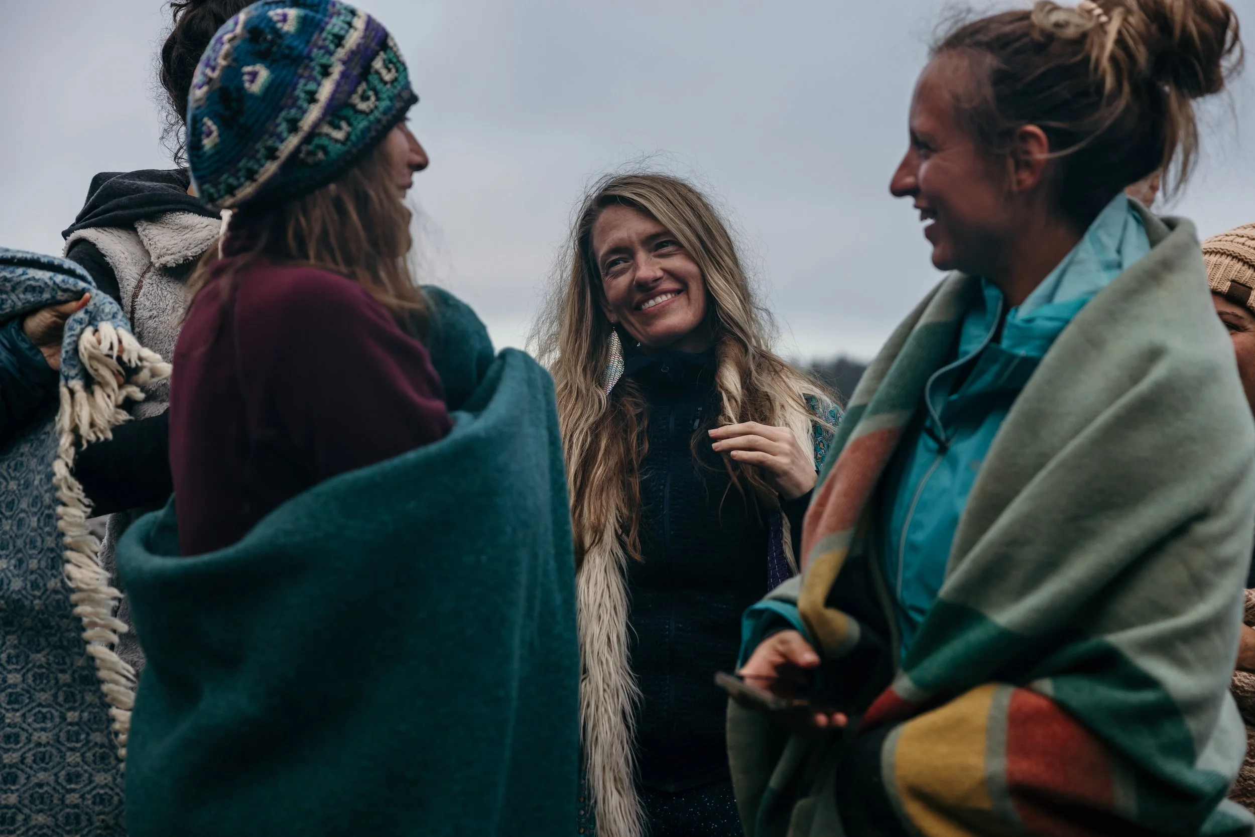 A group of four women smiling and talking outdoors on a cloudy day, dressed in casual jackets and sweaters.