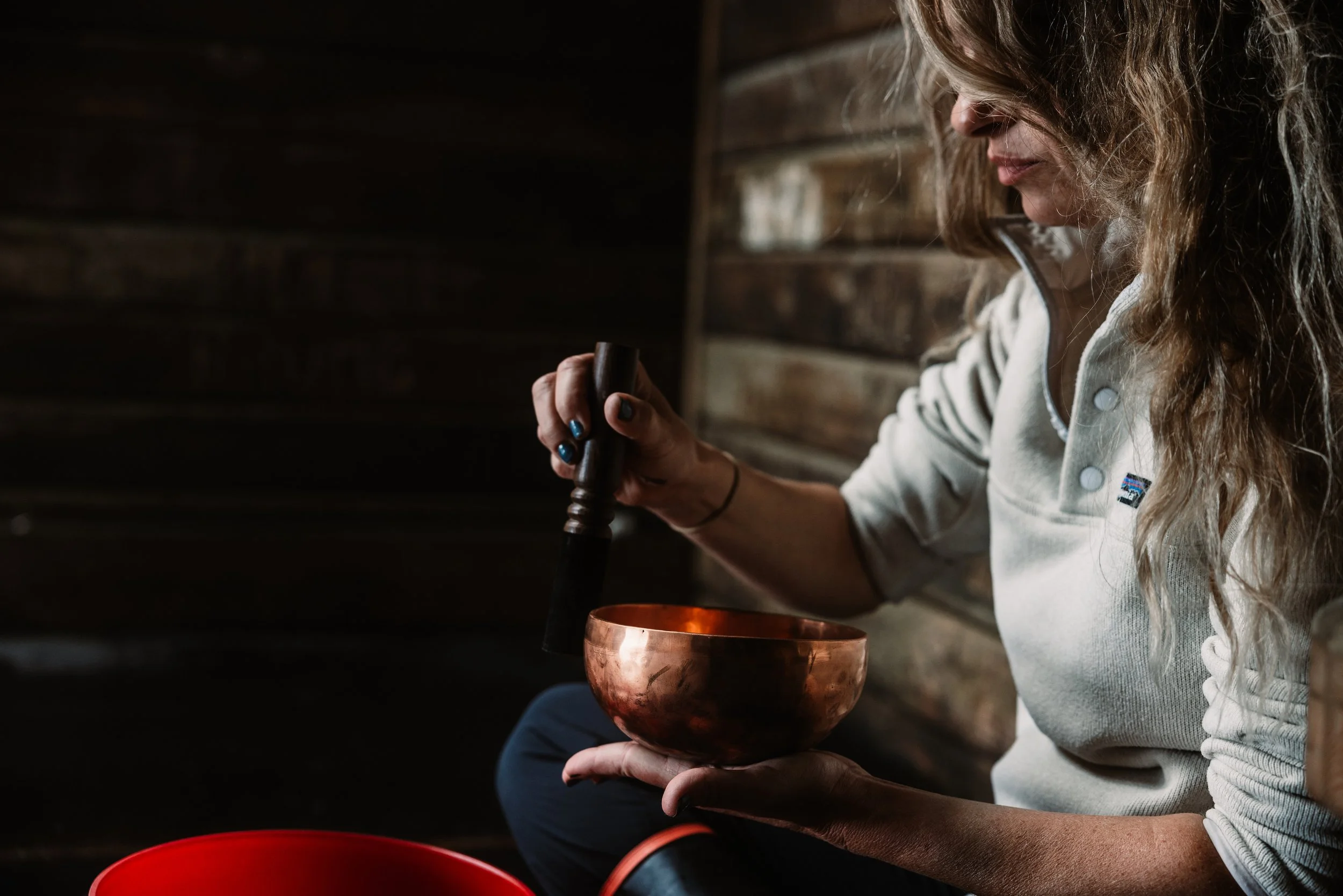 A woman with wavy hair wearing a light gray cardigan is sitting and holding a wooden singing bowl in her lap, striking it with a black mallet. The setting has dark wooden walls.