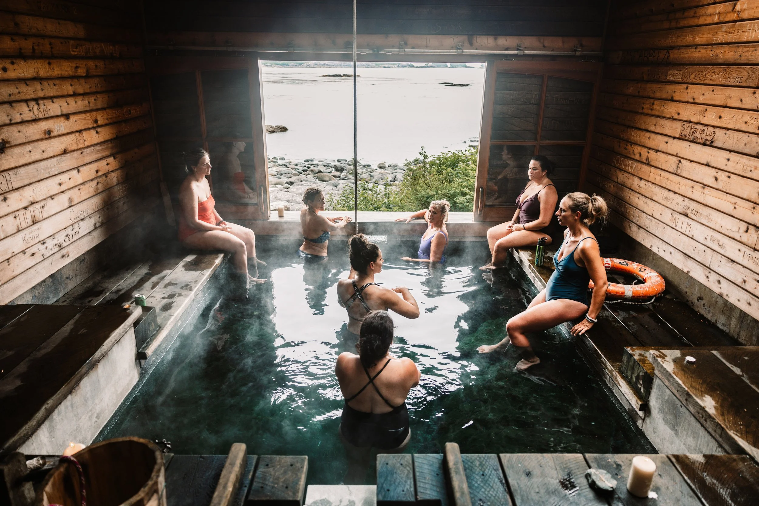 A group of women sitting and standing in an outdoor hot tub with a view of the rocky shoreline and water outside. The wooden interior of the hot tub area is visible.