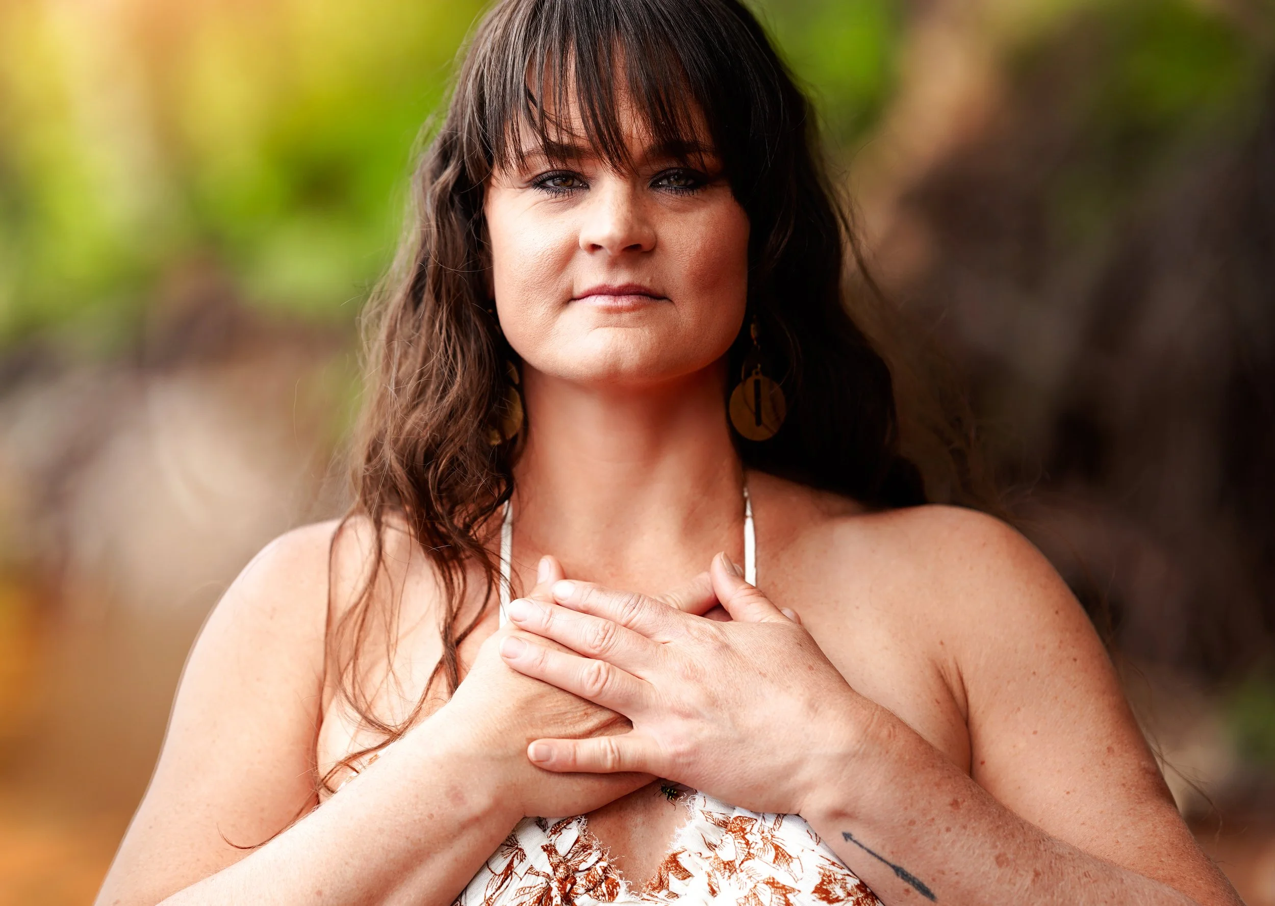 A woman with long brown hair, wearing a white and brown floral dress, is holding her chest with both hands in an outdoor setting. The background is blurred with green and yellow tones.