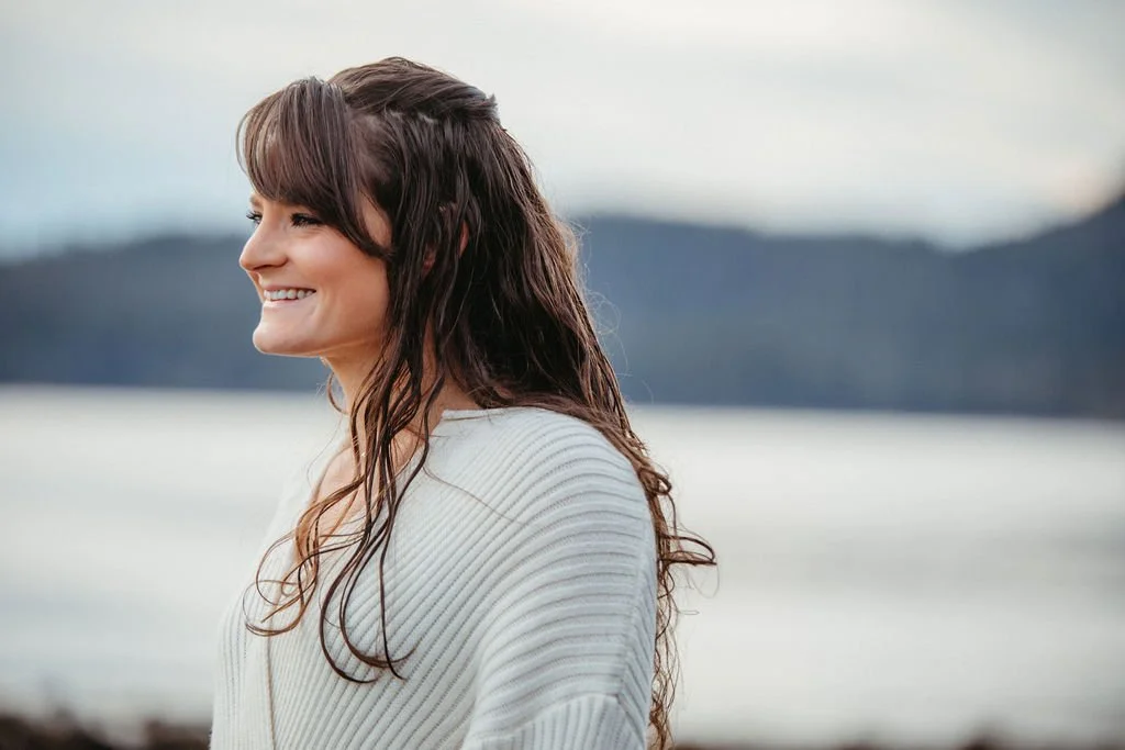 A woman with long wavy brown hair smiling and looking to the side outdoors near a body of water with mountains in the background.