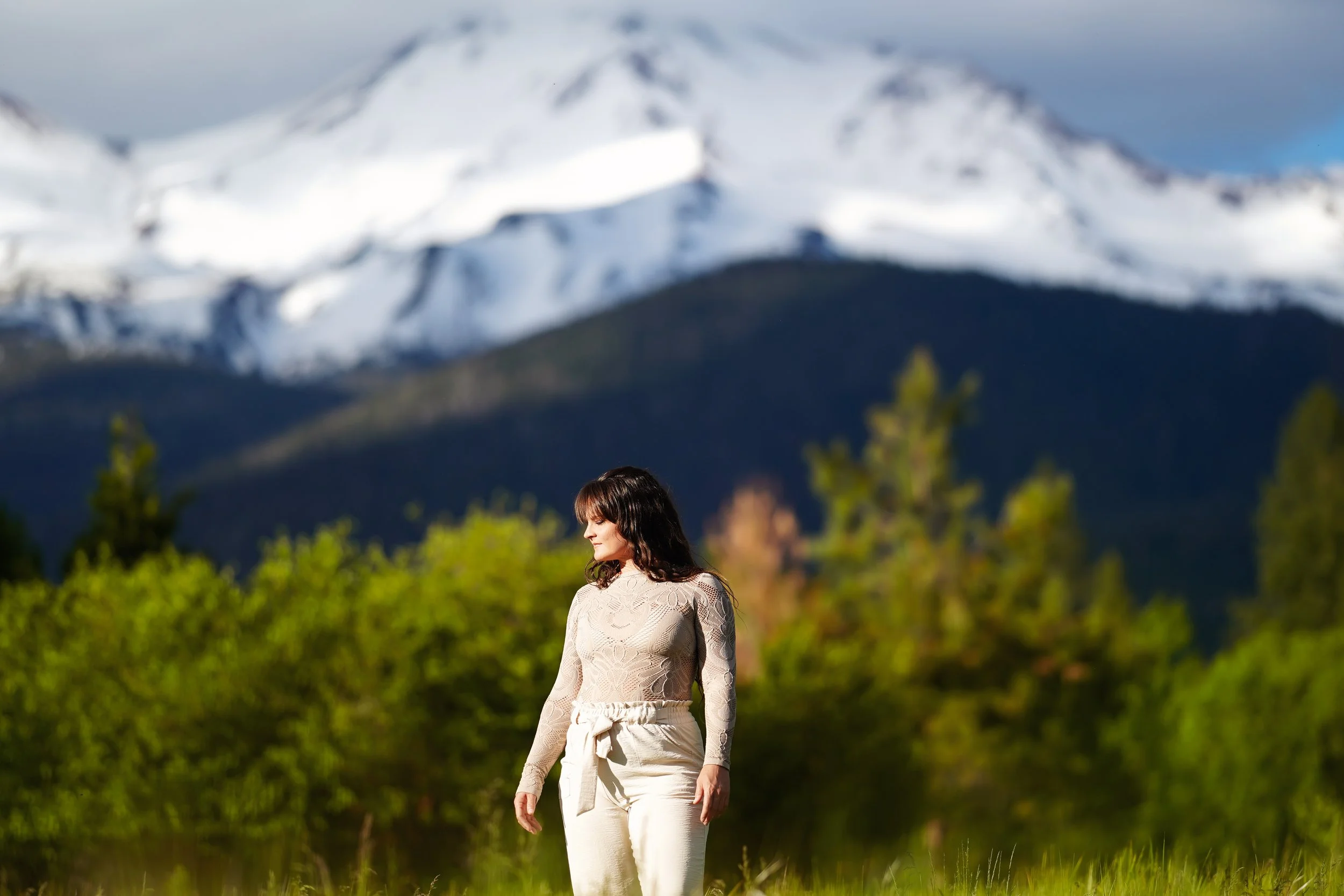 A woman with dark hair wearing a beige lace long-sleeve top and cream pants with a bow, standing outdoors in a green field with snow-capped mountains in the background.