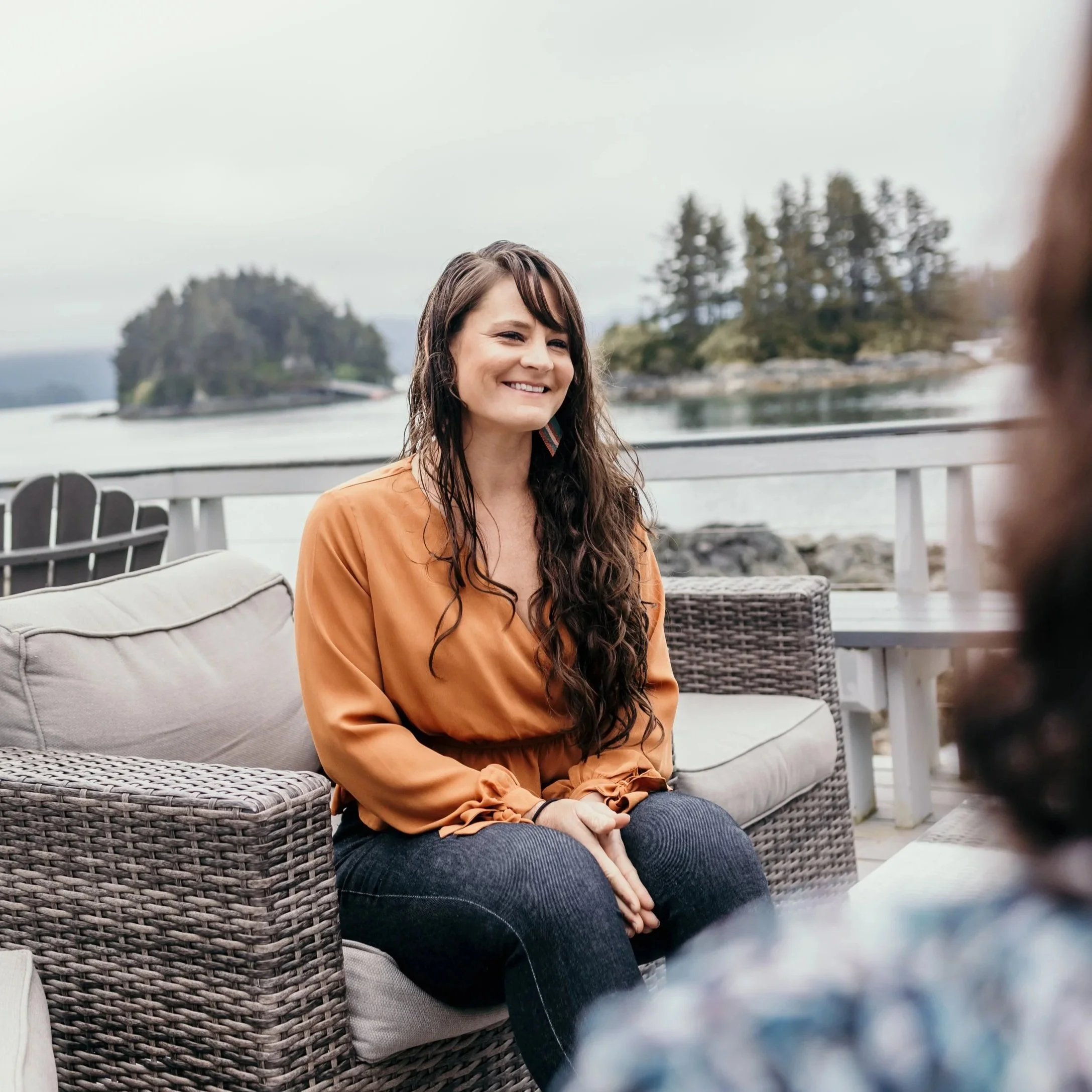 A woman with long wavy hair smiling, sitting outdoors on a wicker chair near water with small islands and trees in the background, overcast sky.