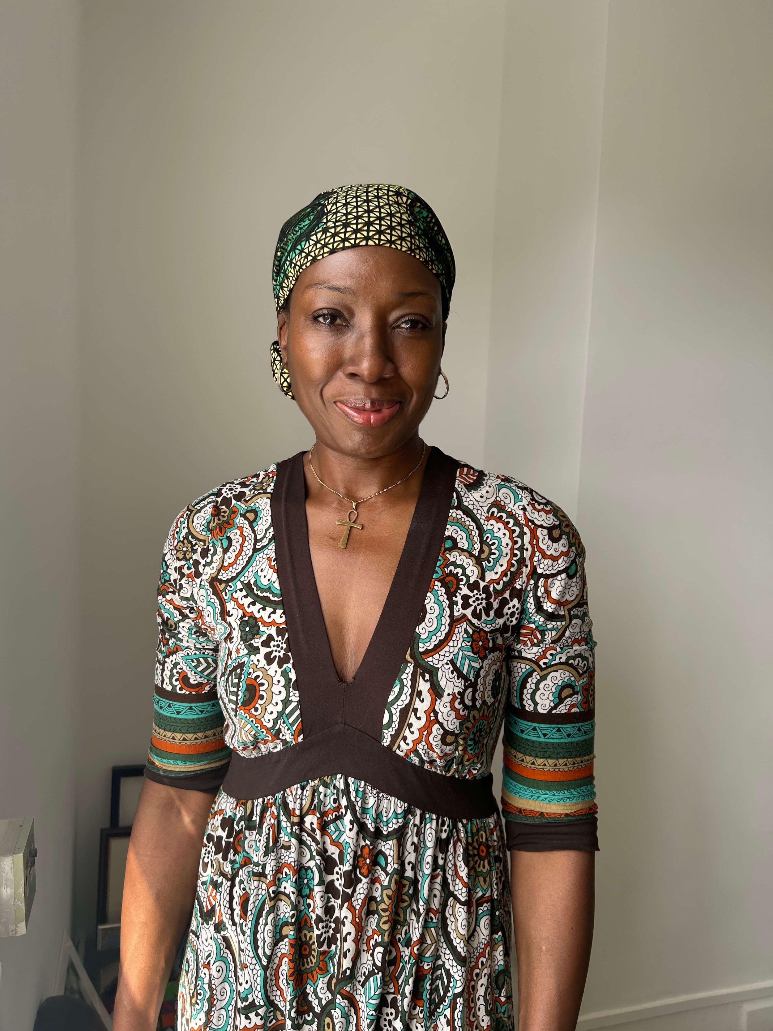 Florence wearing a colorful patterned dress and matching headwrap, with a cross necklace, standing in front of a plain white wall.