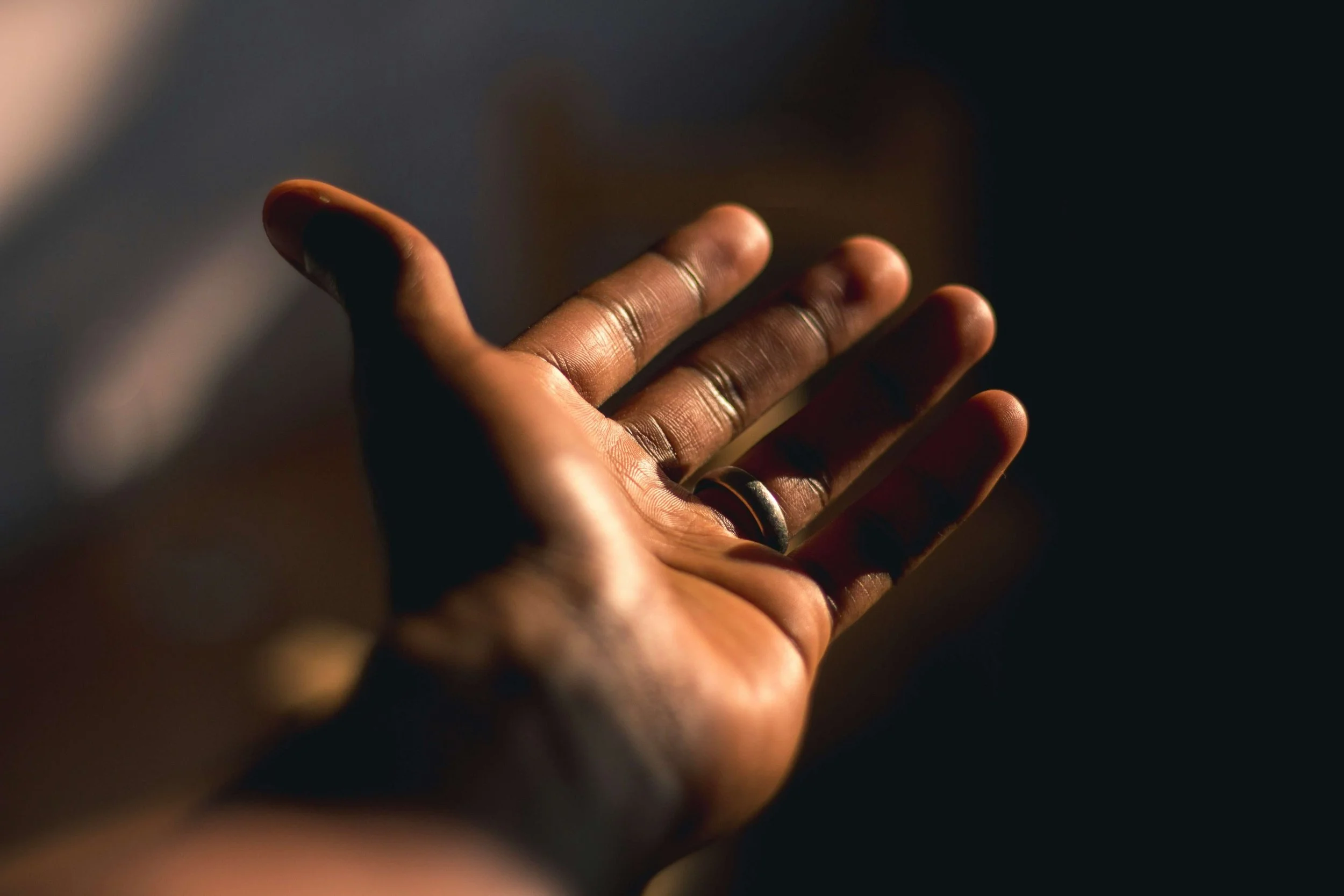Close-up of a person's hand with dark skin, reaching out or gesturing in a dark setting, illuminated softly from one side, with focus on fingers and ring on the finger.