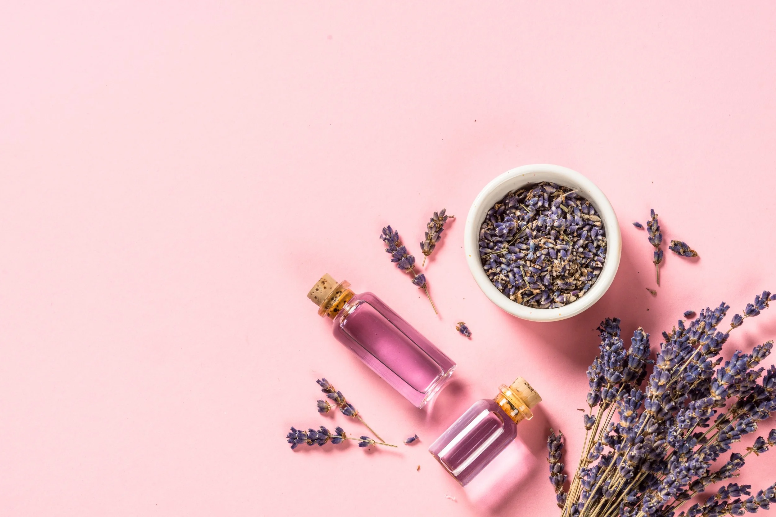 A flat lay of small glass bottles with pink liquid, a white bowl filled with dried lavender, and sprigs of lavender on a pink background.