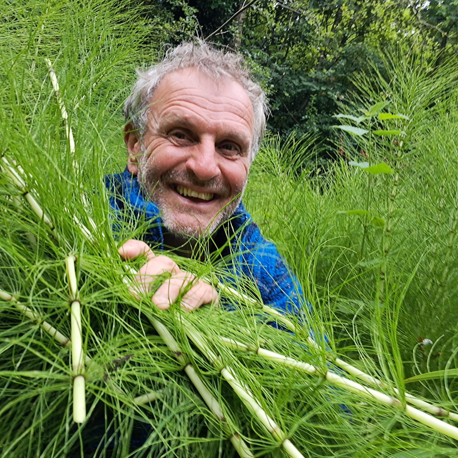 Profile picture of Greg (Touches Sky) plant spirit healer, wearing a light blue jumper surrounded by plants in nature