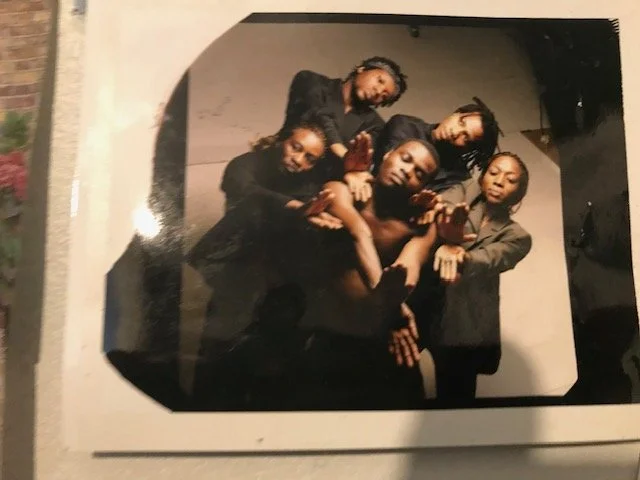 A group of two African women and three African men posing together from former Adzido dance company , making hand gestures, and looking at the camera in a studio setting.