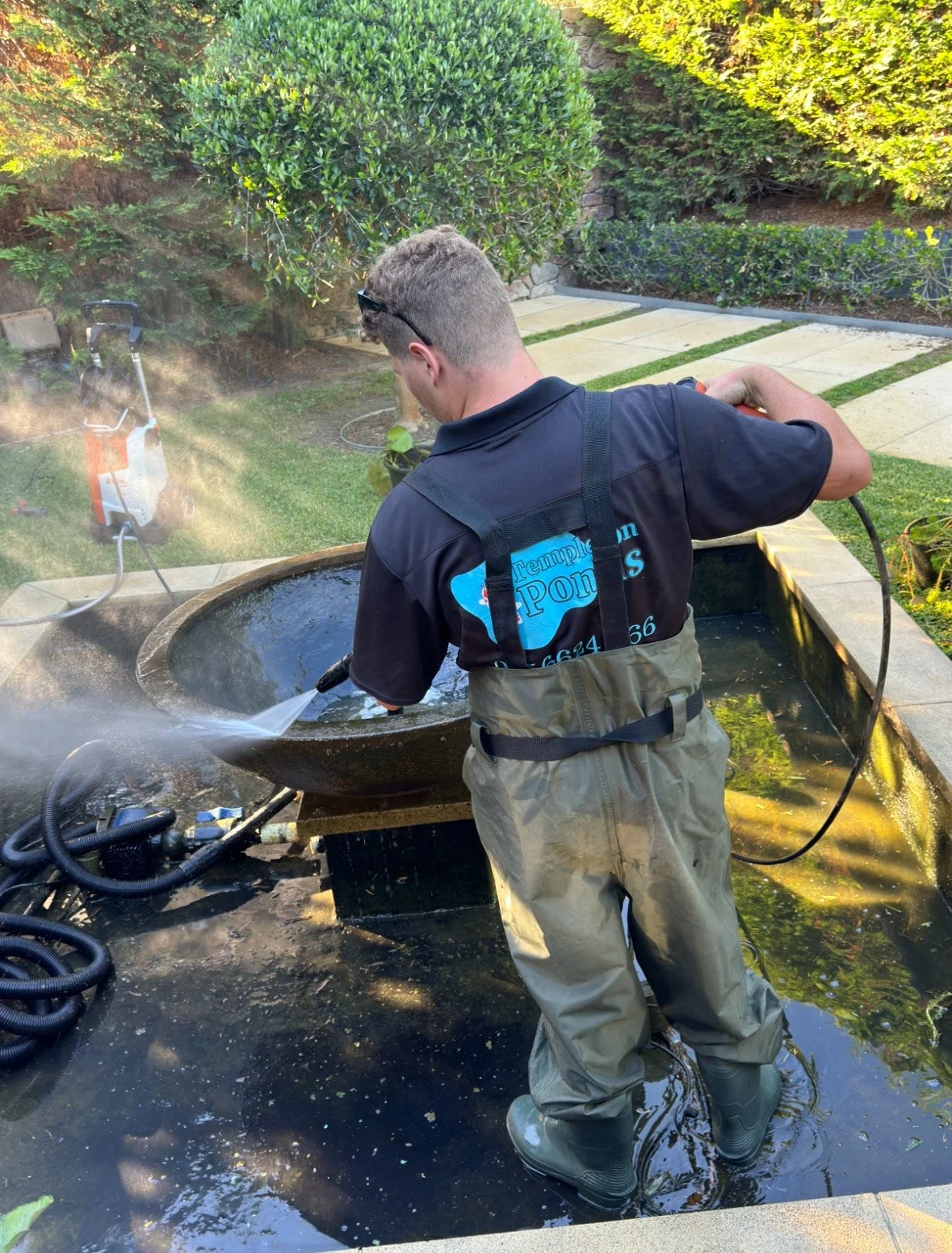 A man wearing waterproof waders and a uniform washing a large pond in a garden. The man is using a pressure washer to clean the pond's interior, with water spraying onto the pond's walls. The garden has green bushes, trees, a stone pathway, and a grassy area.