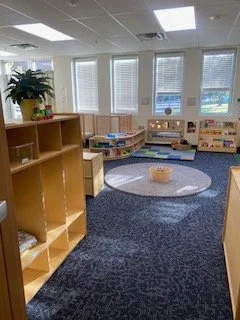 A classroom with large windows, a blue carpet, a bookshelf, and a circular rug with a basket in the center.