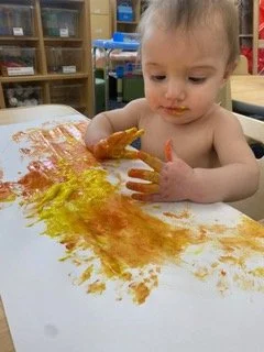 A young child with short hair is sitting at a table, engaging in finger painting with orange, yellow, and red paint, on a sheet of paper.