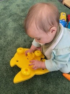 Infant playing on a yellow activity mat with various attached toys, sitting on green carpet.