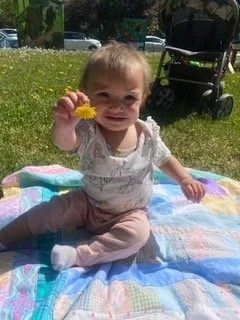 A smiling baby girl sitting on a blanket outdoors, holding a yellow flower, with a stroller in the background.