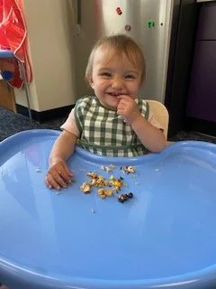 A smiling young child with light brown hair sitting in a blue high chair, wearing a green checkered smock, with food on the tray in front of her, in a kitchen setting.