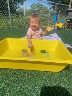 A young girl playing in a yellow sandbox outdoors, holding a yellow toy, with a playground and fence in the background.