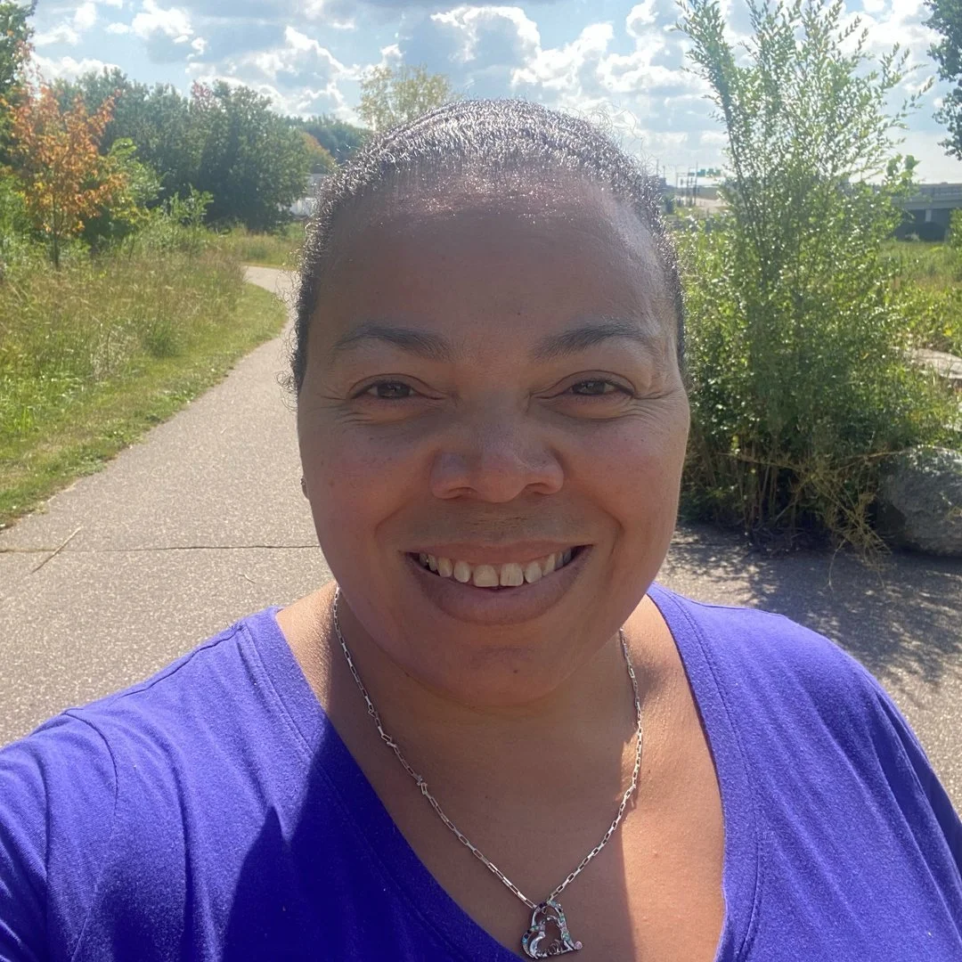 A woman smiling outdoors on a sunny day, wearing a purple shirt and a heart-shaped necklace, with a winding path, trees, and blue sky in the background.