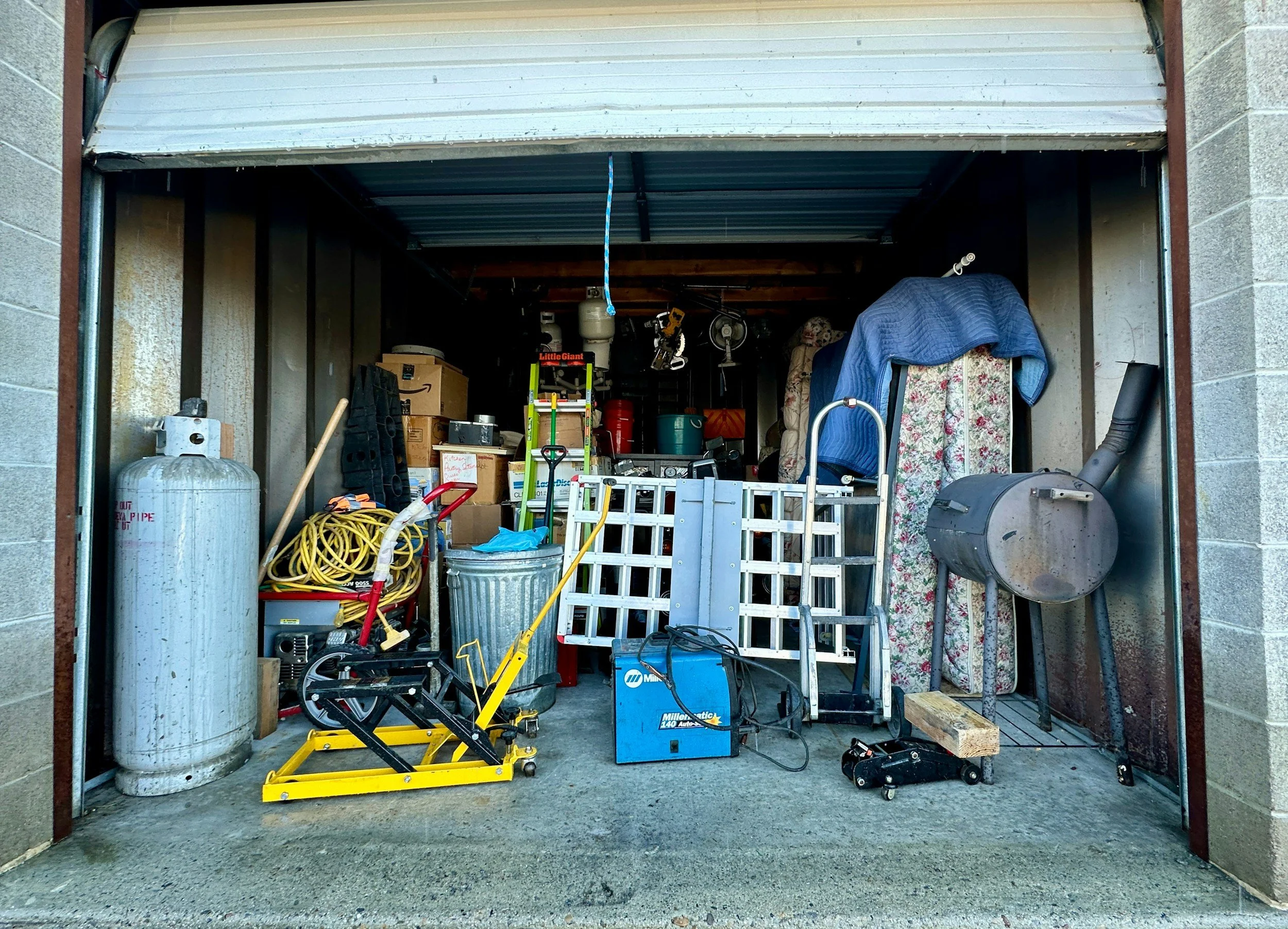 The image shows an open garage filled with various tools, equipment, and stored items. Items visible include a propane tank, a yellow mechanical lift, a blue Miller welder, a garden hose, boxes, a ladder, and other miscellaneous household and workshop items.