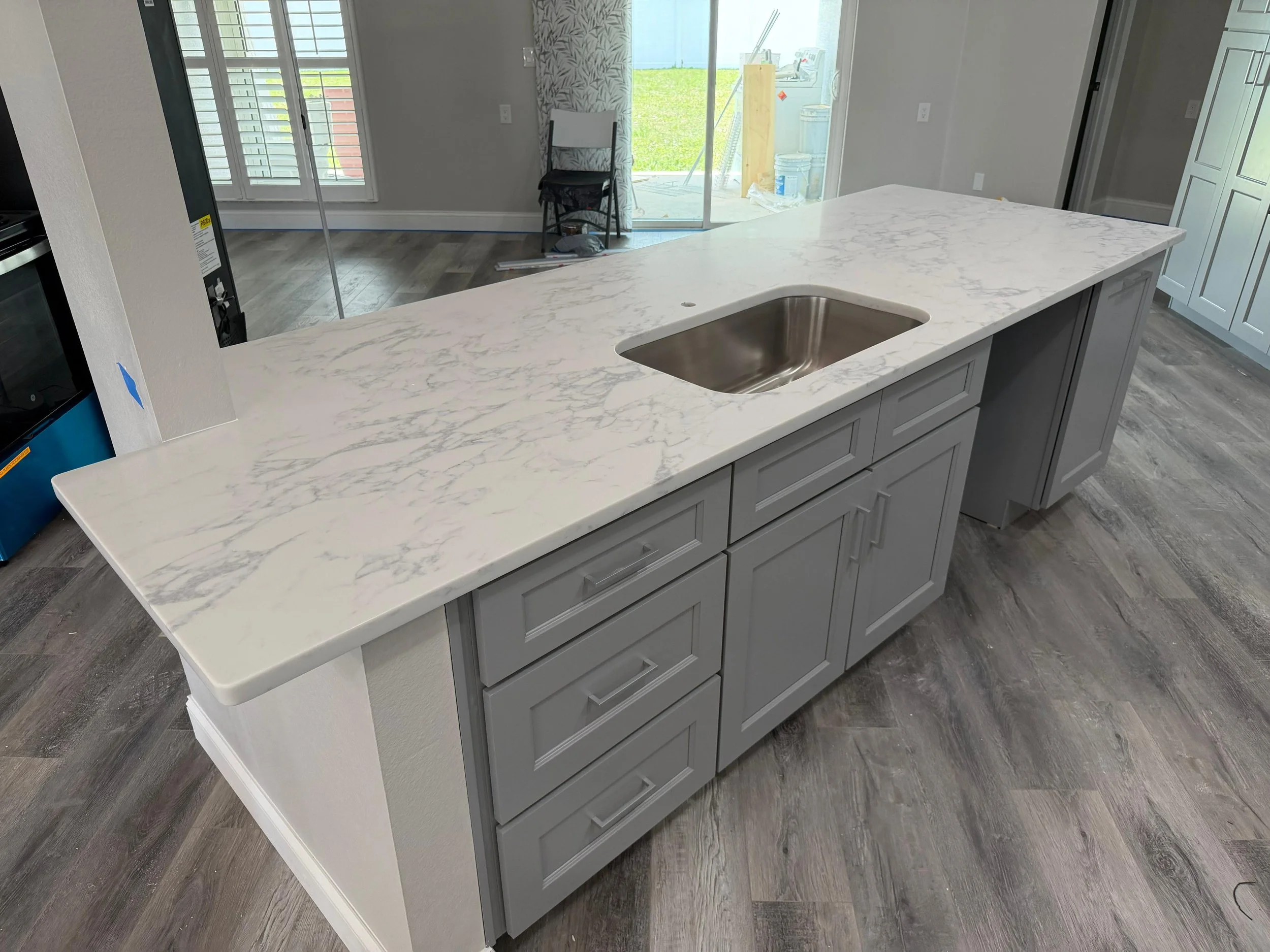 Kitchen island with white marble countertop and gray cabinetry, stainless steel sink, in a modern kitchen with gray flooring and light-colored walls, sliding glass door leading to a backyard.
