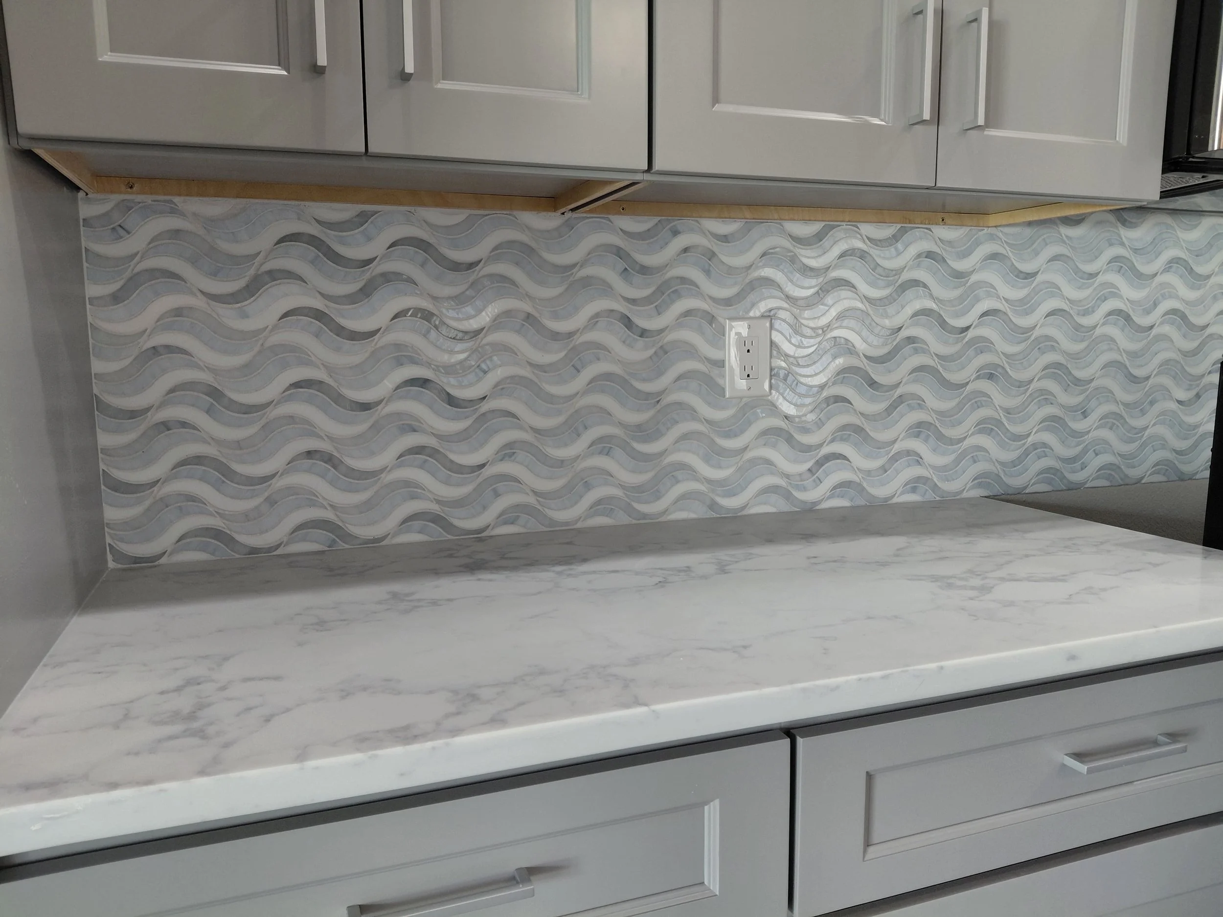 Close-up of a kitchen countertop with a marble pattern, white cabinet drawers below, and a decorative backsplash with wavy gray and white tiles. An electrical outlet is visible on the backsplash.