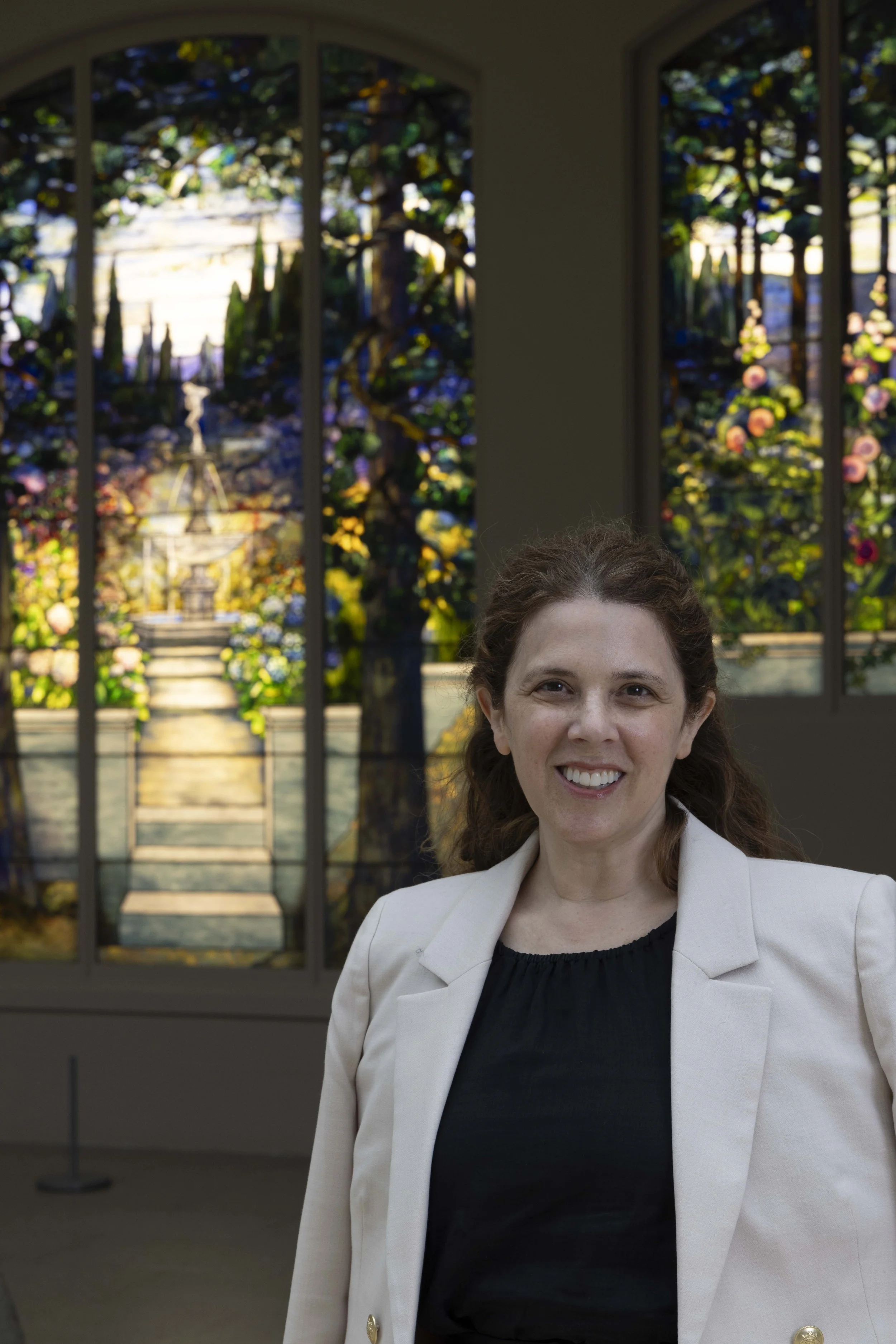 A woman with brown hair wearing a black top and a beige blazer, standing inside a building with large stained-glass windows depicting a garden scene, smiling at the camera.