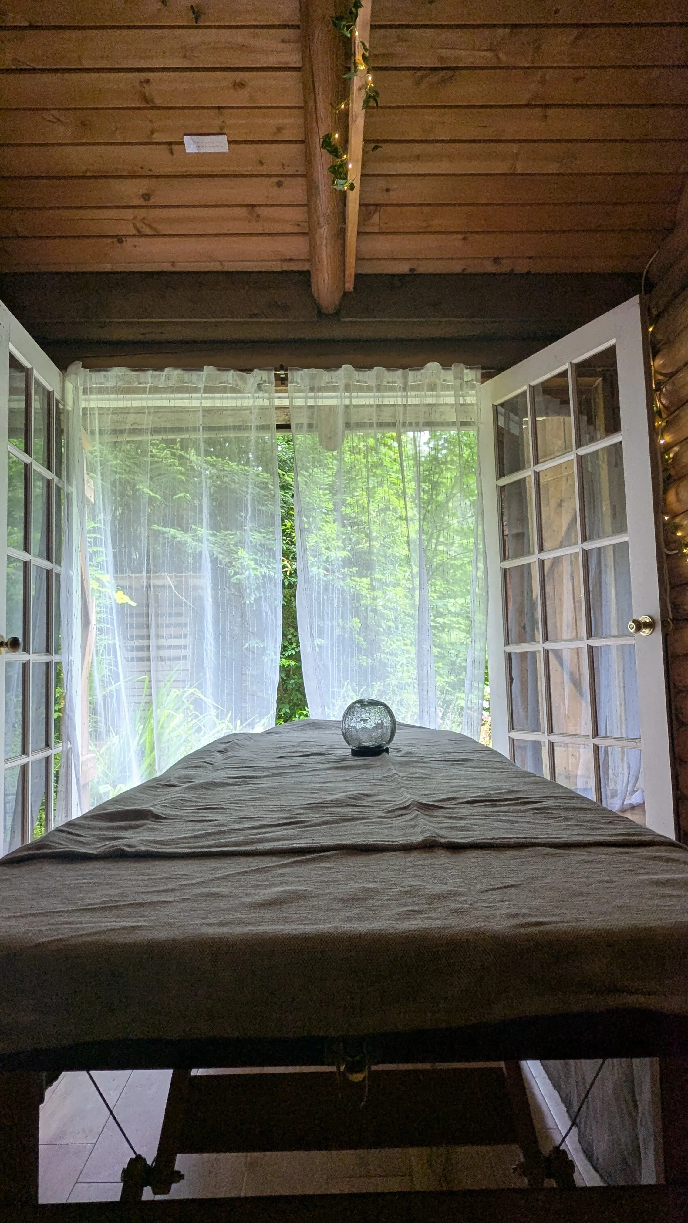 Open window with white curtains revealing greenery outside, with a round glass candle holder on a bed or table inside, and a wooden ceiling with string lights.