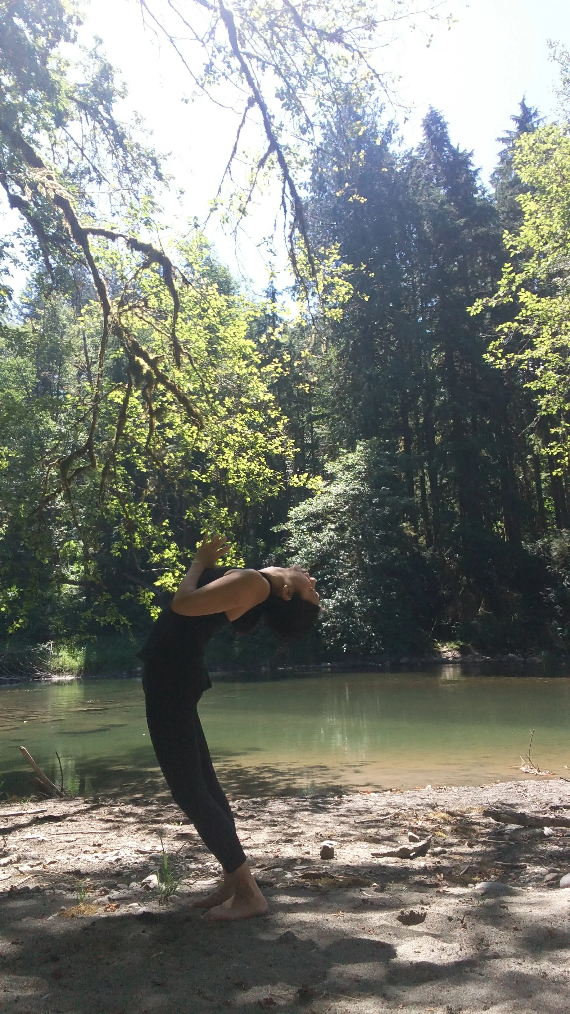 A person practicing yoga in a backbend pose on a sandy riverbank surrounded by trees and water.