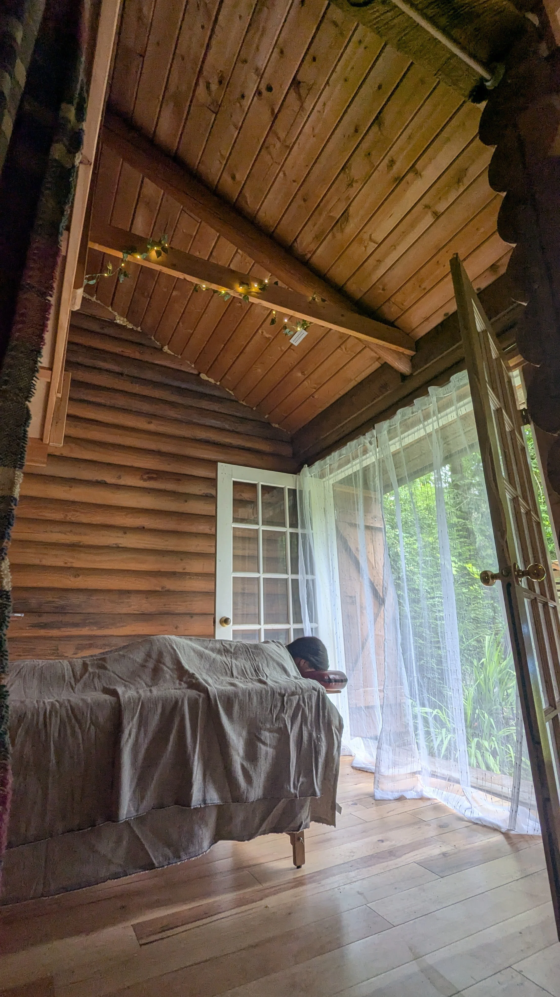 Interior of a rustic cabin room with wood-paneled ceiling and walls, a window with sheer curtains, a couch covered with a beige sheet, and a person resting their head on the armrest.