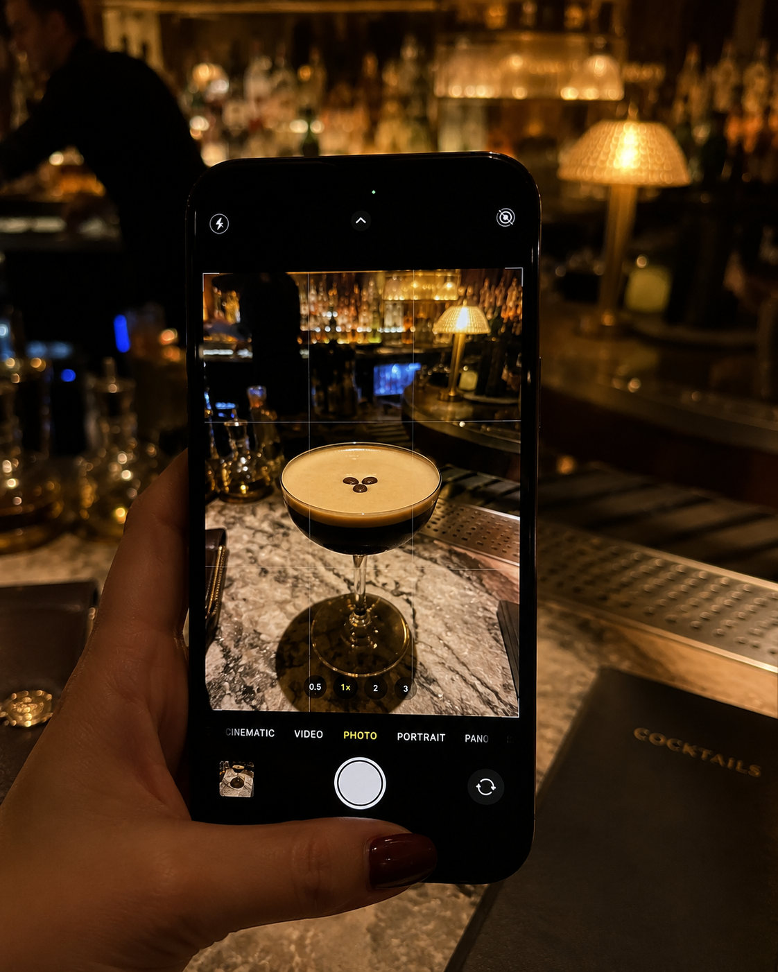 A person is taking a photo of a dark coffee cocktail with three coffee beans on top, in a cocktail glass at a bar, with a bar counter, bottles, a lamp, and a bartender in the background.